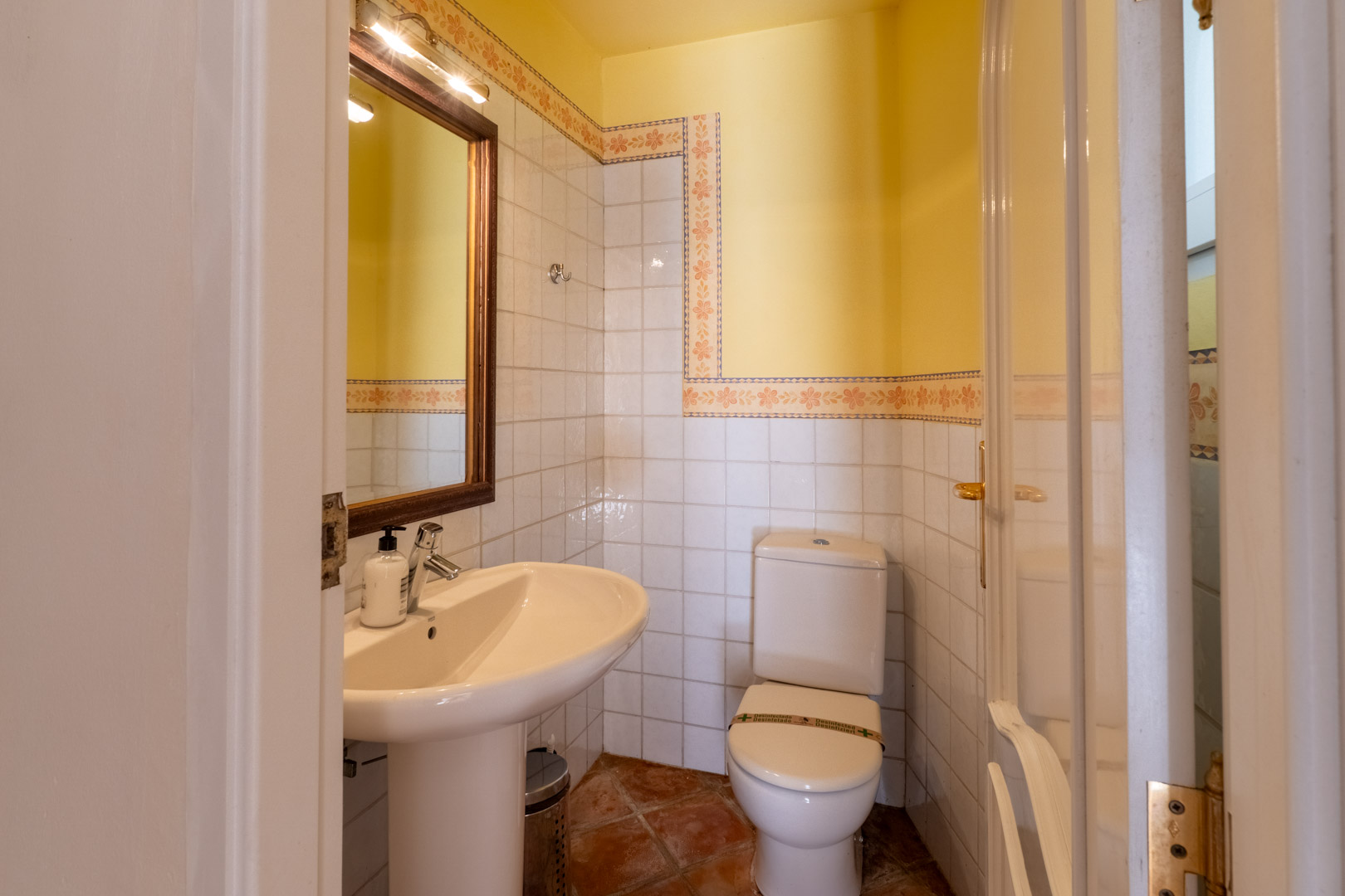 Gäste-WC mit Terrakottaboden, weiß gefliesten Wänden mit floraler Bordüre und einem Säulenwaschbecken unter einem beleuchteten Holzrahmenspiegel.