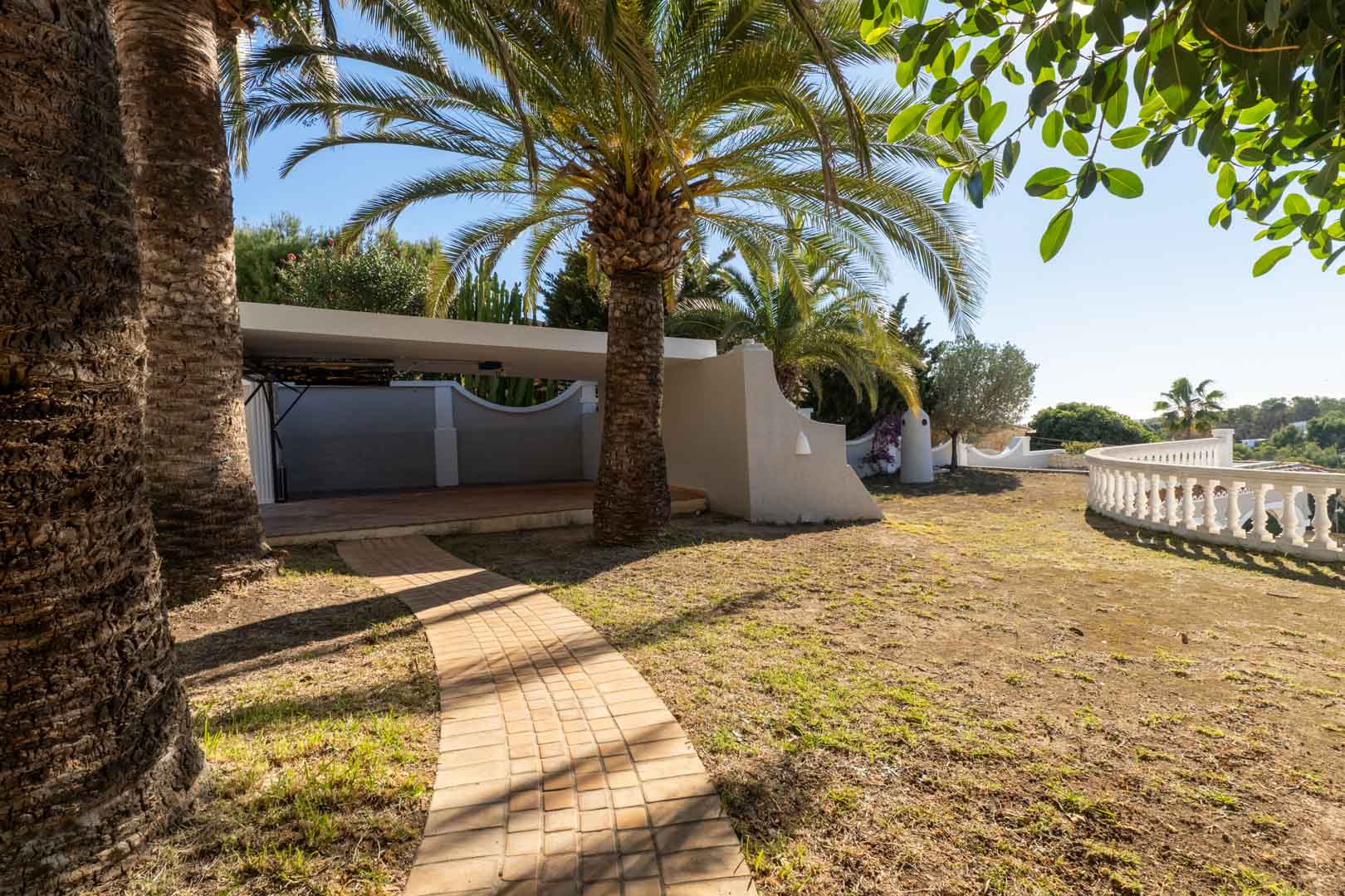 Gepflasterter Weg, der zu einem überdachten Carport führt, gesäumt von hohen Palmen und einer weißen Balustrade mit weitem Ausblick.