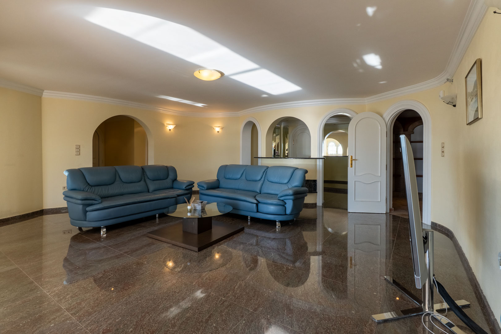 Großzügiger Wohnraum mit glänzendem Marmorboden, blauen Ledersofas und Rundbögen, die zu einer Bar und angrenzenden Räumen führen.