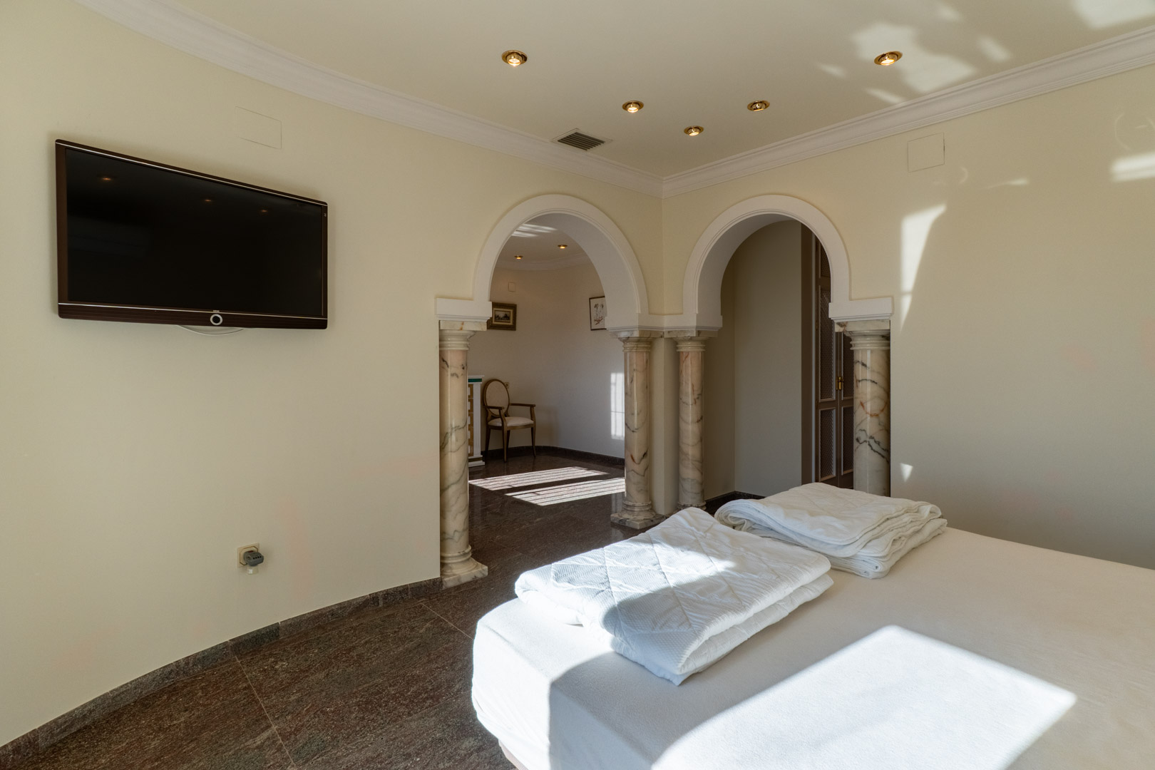 Hauptschlafzimmer mit klassischen Rundbögen auf Marmorsäulen, poliertem Natursteinboden und direktem Zugang zu einem hellen angrenzenden Bereich.
