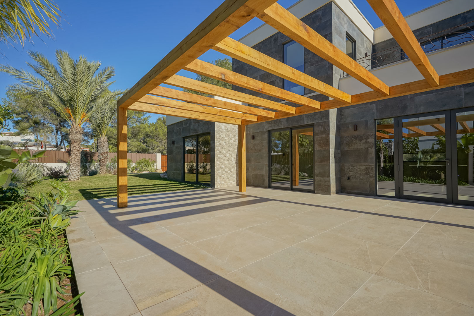 Außen-Terrasse mit einer Pergola aus Iroko-Holz, großformatigen Bodenfliesen und einer Fassade aus Natursteinmauerwerk im mediterranen Garten.