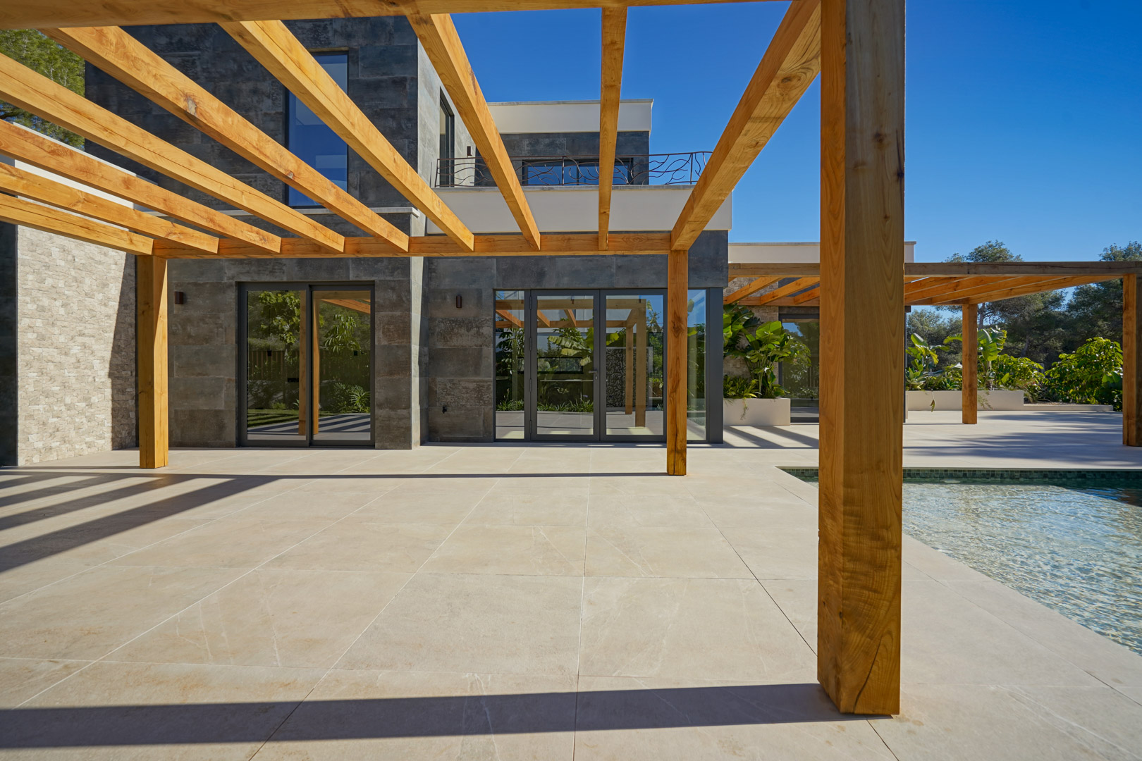 Außen-Terrasse mit Iroko-Holzpergola und hellen Fliesen, angrenzend an den Pool und eine moderne Fassade mit großen Glasfronten.