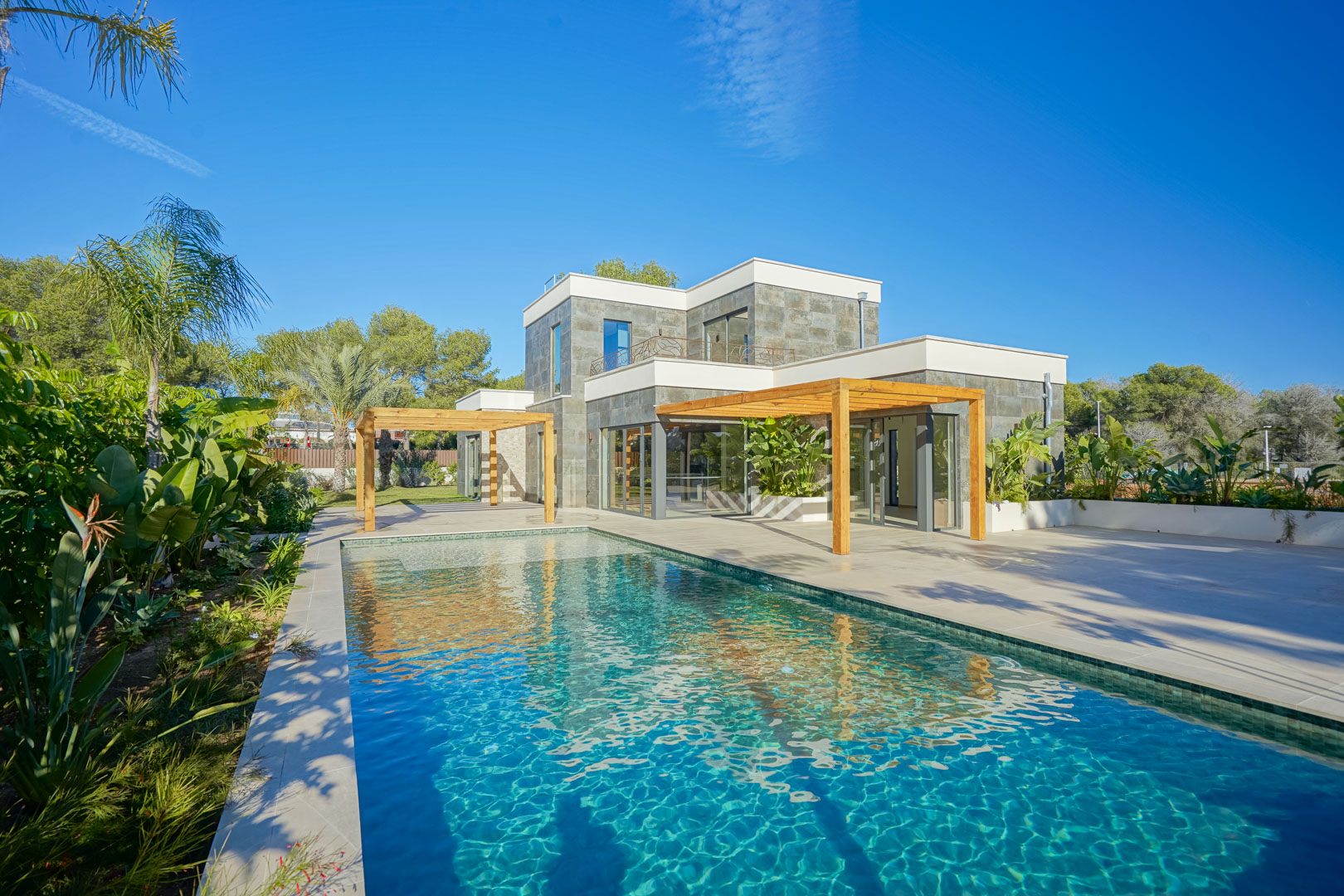 Außenansicht der Villa mit Pool im Bali-Stil, großzügigen Terrassen und Pergolen aus Iroko-Holz vor der modernen Steinfassade.