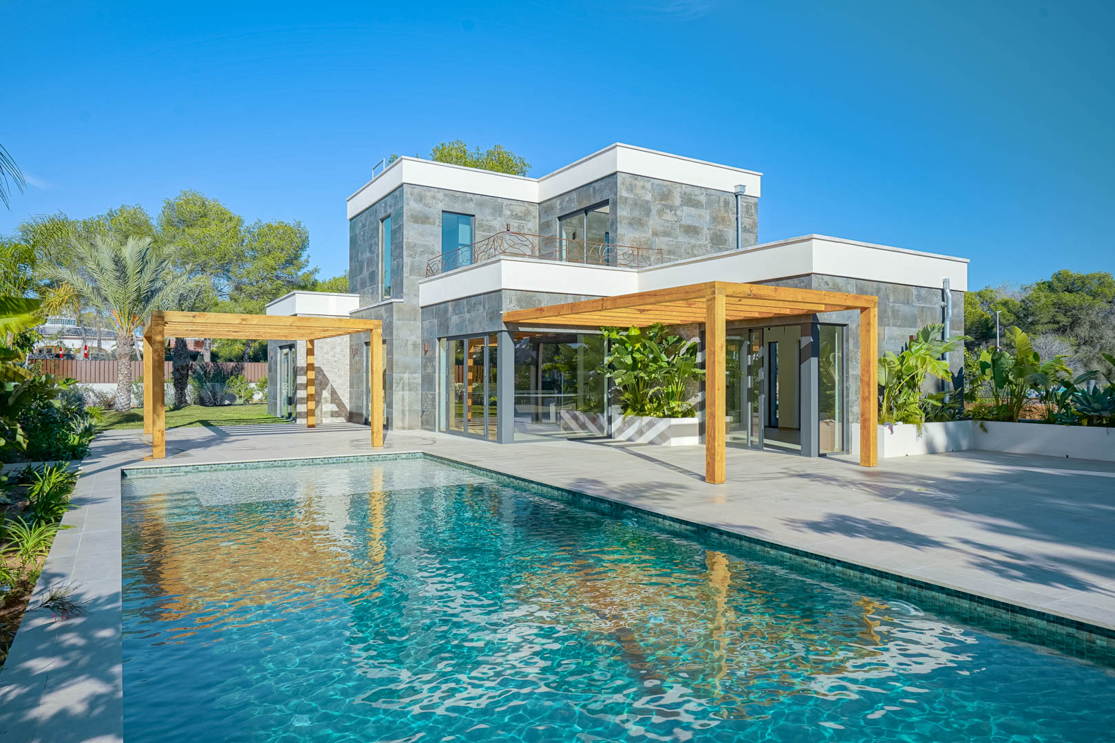 Außenansicht der Villa mit Pool im Bali-Stil, weitläufigen Terrassen und Iroko-Holzpergolen an einer modernen Fassade mit Natursteinverkleidung.