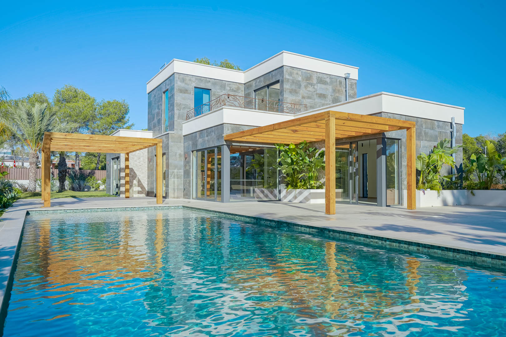 Außenansicht der Villa mit türkisfarbenem Mosaikpool, Pergolen aus Iroko-Holz und einer Natursteinfassade mit großen Glasfronten.