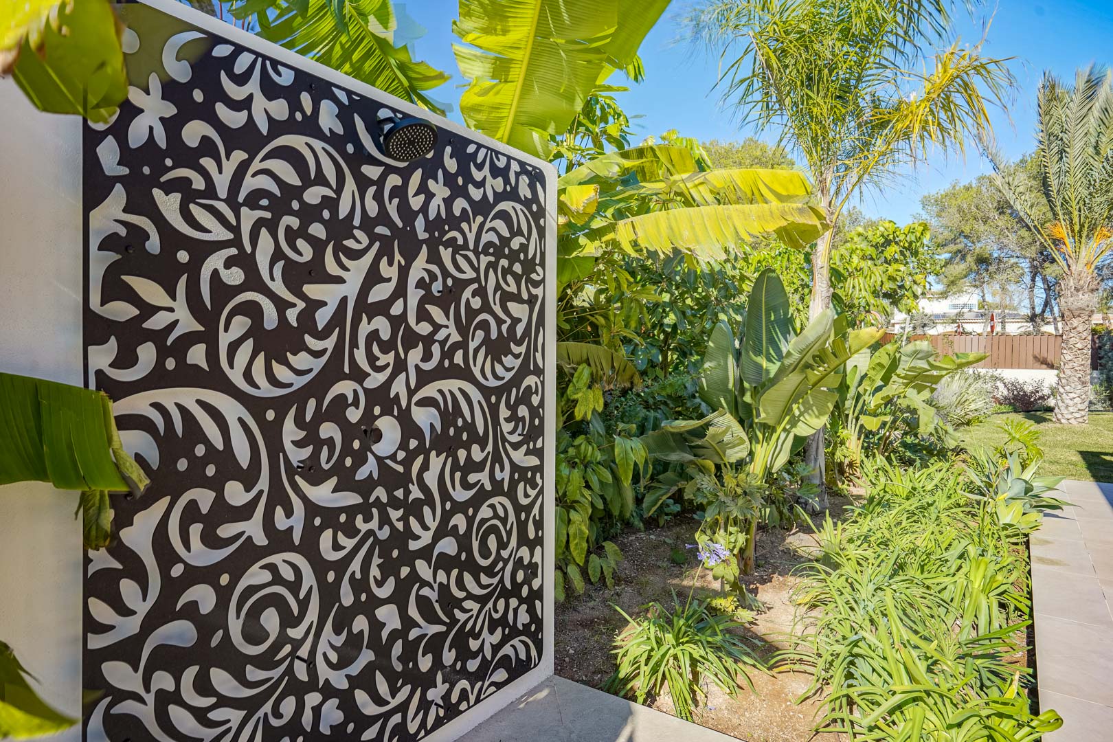 Außendusche mit dekorativem, lasergeschnittenem Paneel und schwarzen Armaturen, eingebettet in einen üppigen mediterranen Garten.