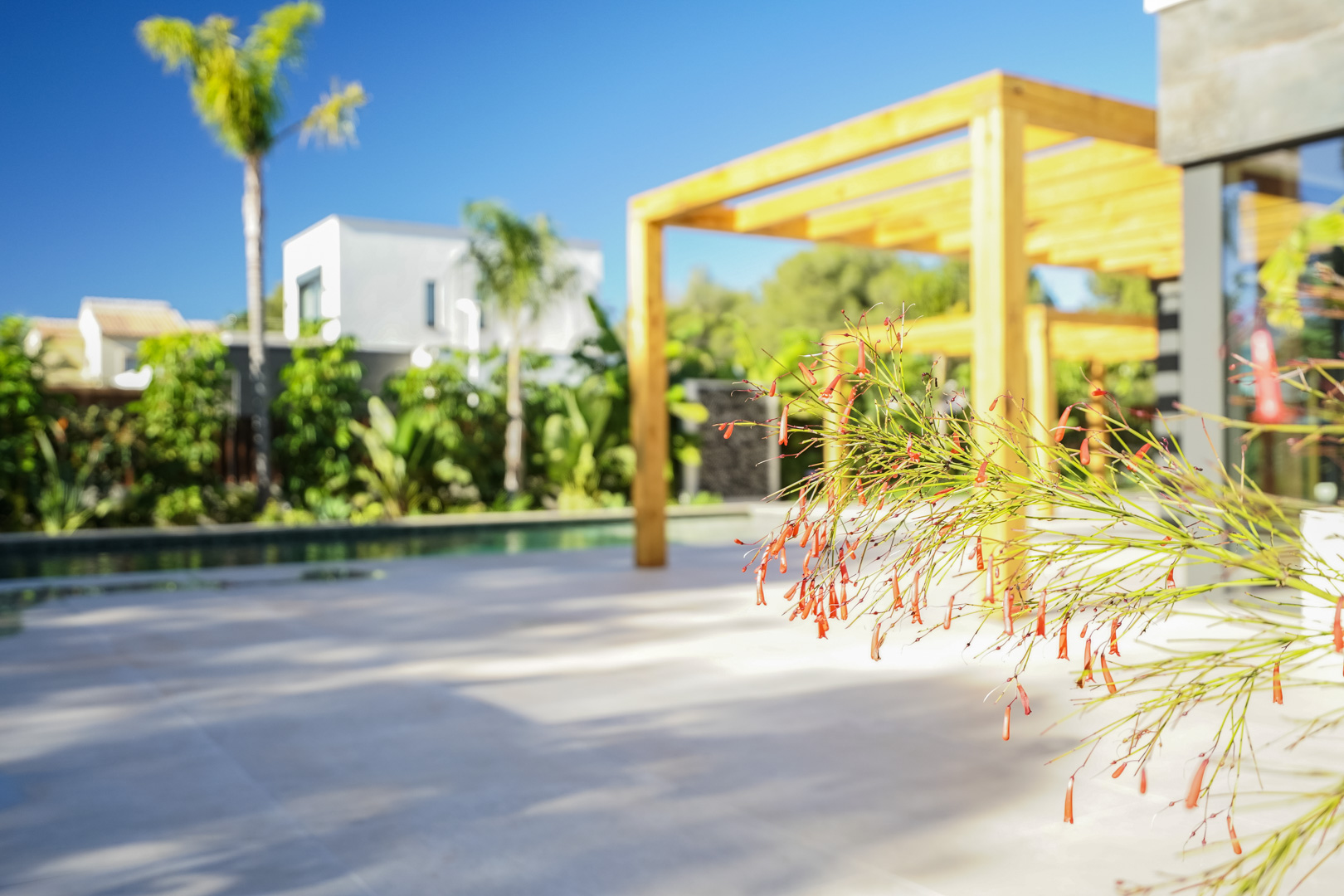 Außenterrasse mit einer Pergola aus Iroko-Holz und Blick auf den mediterranen Garten. Im Vordergrund Zierpflanzen neben dem Poolbereich.