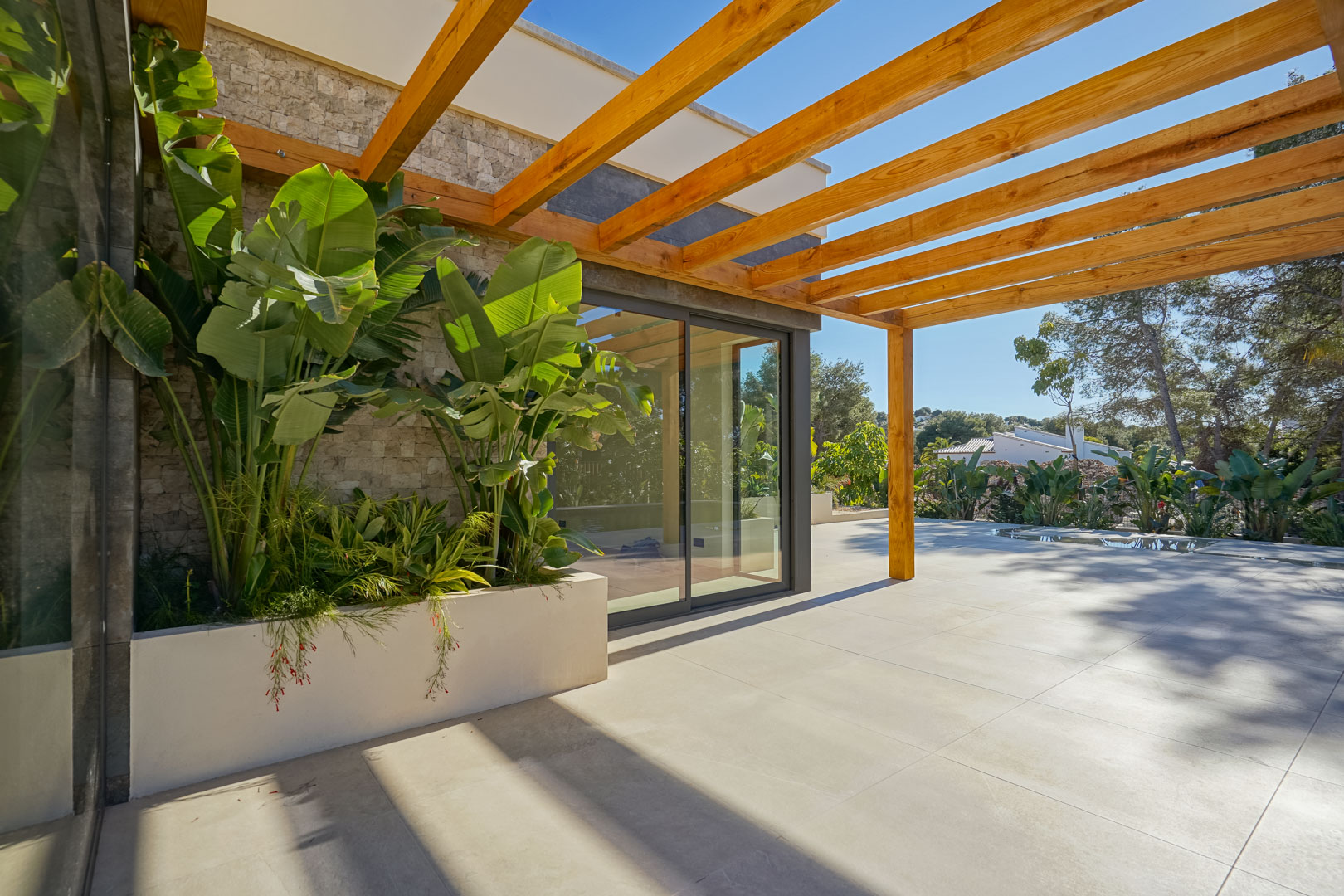 Außenterrasse mit Iroko-Holzpergola, großformatigen Fliesen und integriertem Pflanzgefäß mit tropischen Pflanzen vor der Natursteinfassade.
