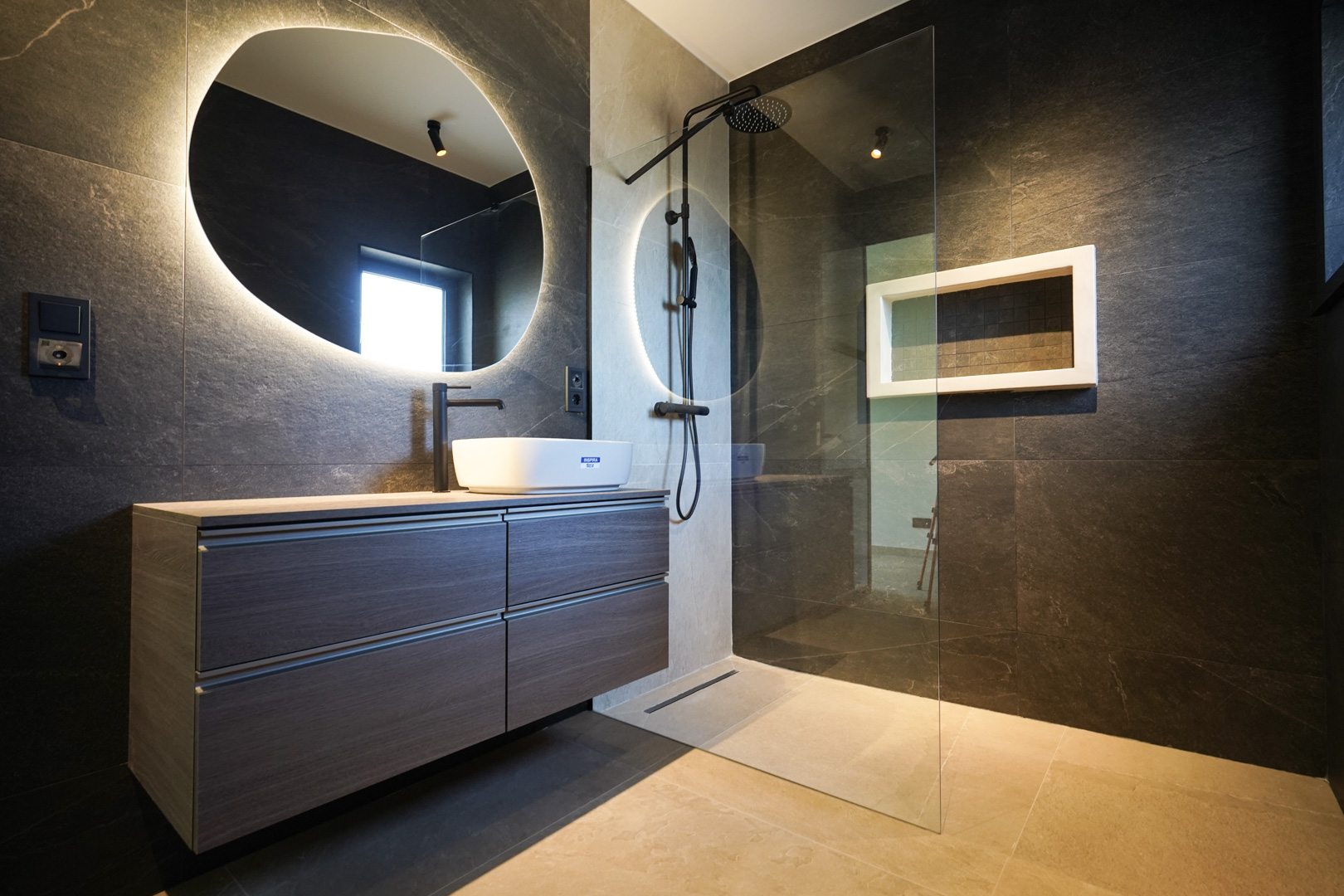 Design-Badezimmer mit dunklen Steinwänden, Aufsatzwaschbecken und hinterleuchtetem organischem Spiegel. Begehbare Dusche mit schwarzen Armaturen und Glastrennwand.