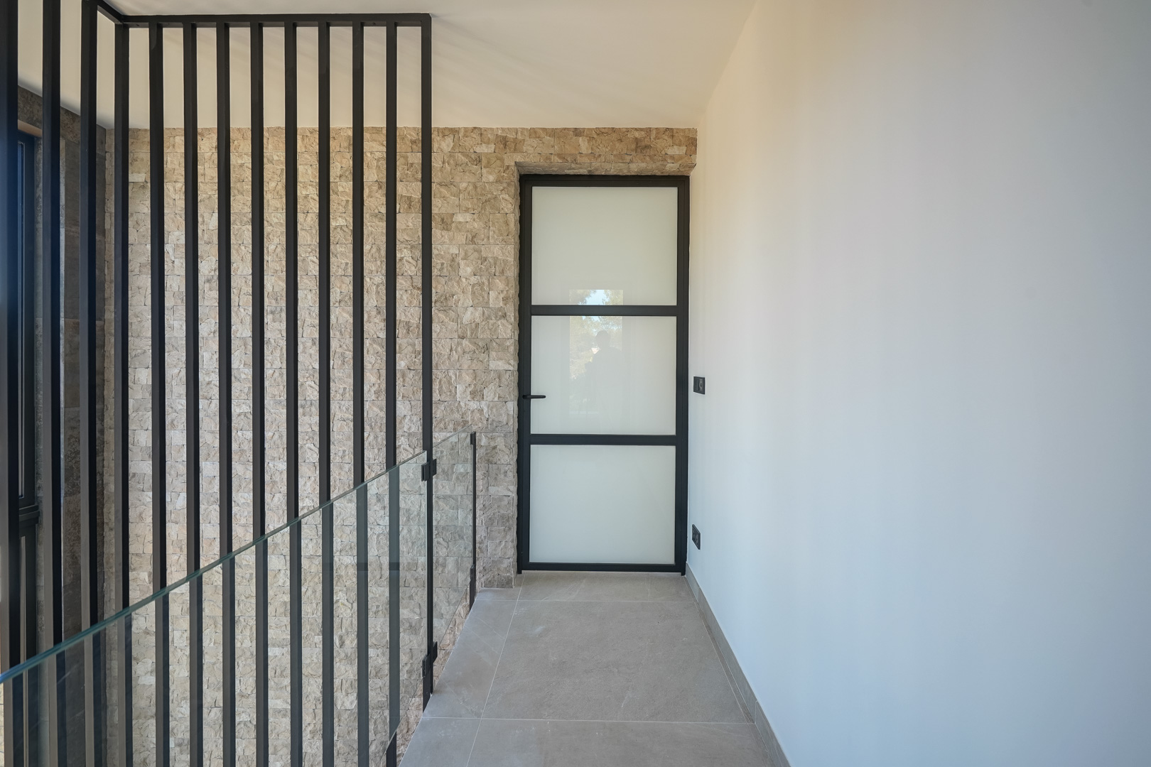 Flur im Obergeschoss mit einer Wand aus Naturstein, Glasgeländer mit schwarzen Metallstreben und einer Industrietür mit mattiertem Glas.