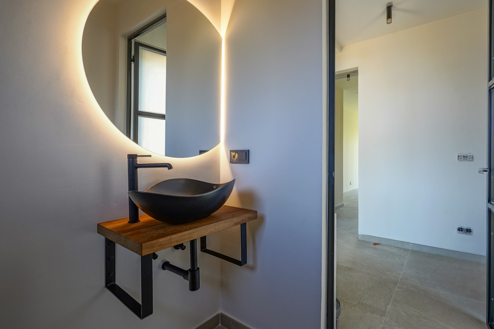 Gäste-WC mit schwarzem Aufsatzwaschbecken auf Massivholzplatte, hinterleuchtetem Rundspiegel und mattschwarzen Armaturen in modernem Design.