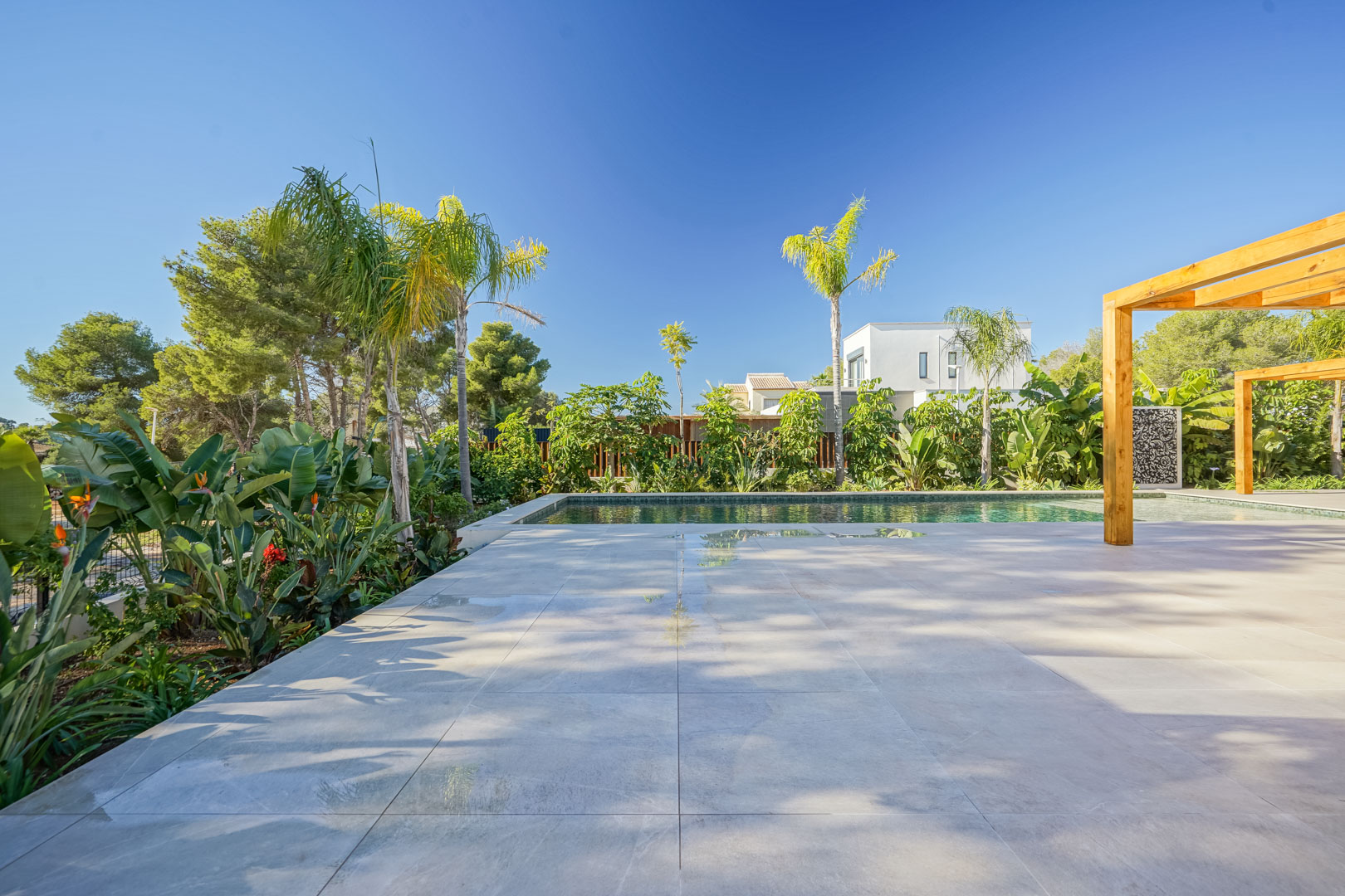 Großzügige Terrasse mit Feinsteinzeugfliesen am Bali-Pool, ergänzt durch Pergolen aus Iroko-Holz und einen üppigen mediterranen Garten.