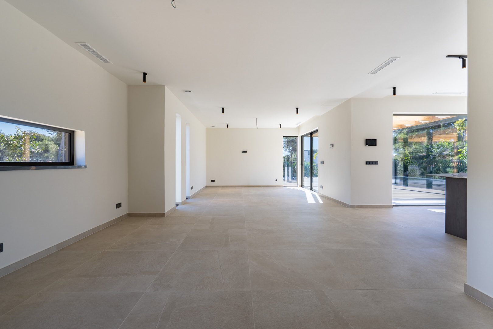 Großzügiger, offener Wohnbereich mit großformatigen Bodenfliesen, minimalistischer Beleuchtung und direktem Zugang zur Terrasse durch Glasschiebetüren.