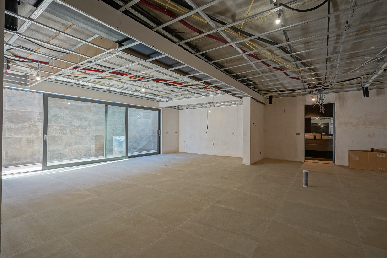 Großzügiges Untergeschoss im Ausbau mit großformatigen Bodenfliesen, Glasschiebetüren und sichtbarer Deckeninstallation für technische Systeme.