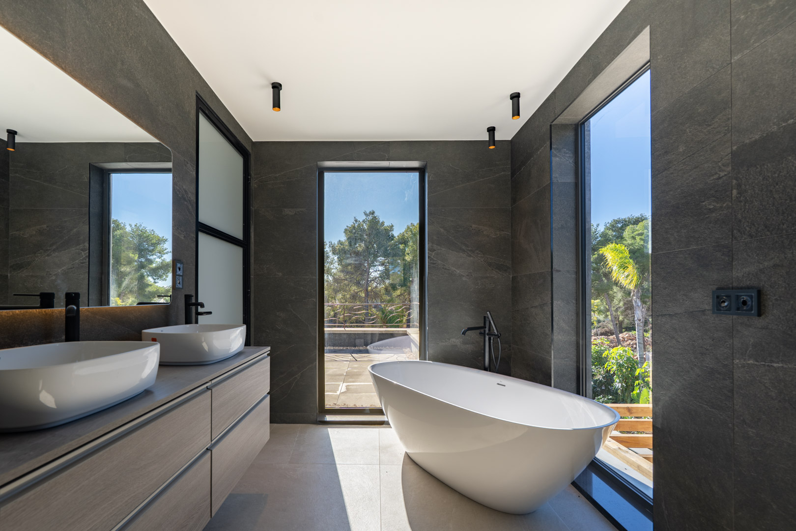 Hauptbadezimmer mit freistehender Badewanne, Doppelwaschtisch und bodentiefen Fenstern mit Blick ins Grüne. Dunkle Steinwände und moderne schwarze Armaturen setzen Akzente.