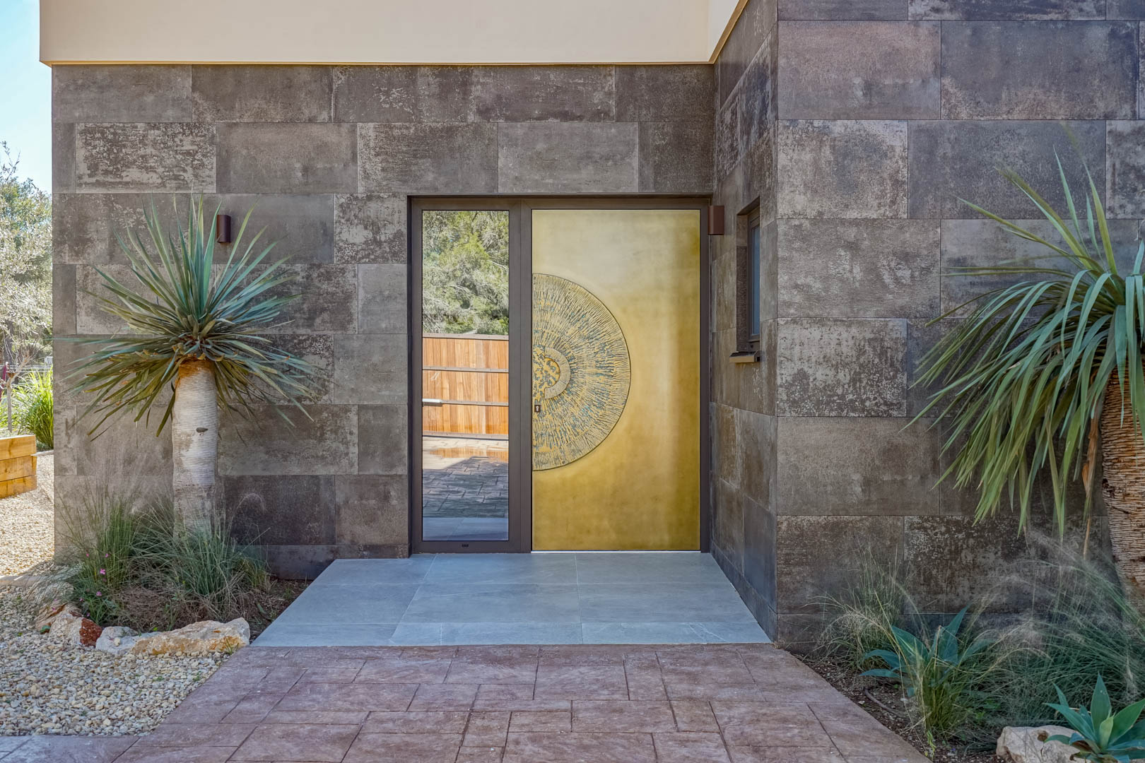Haupteingang mit einer goldenen Designtür mit kreisförmigem Relief, umrahmt von dunklen Natursteinwänden und mediterraner Bepflanzung.