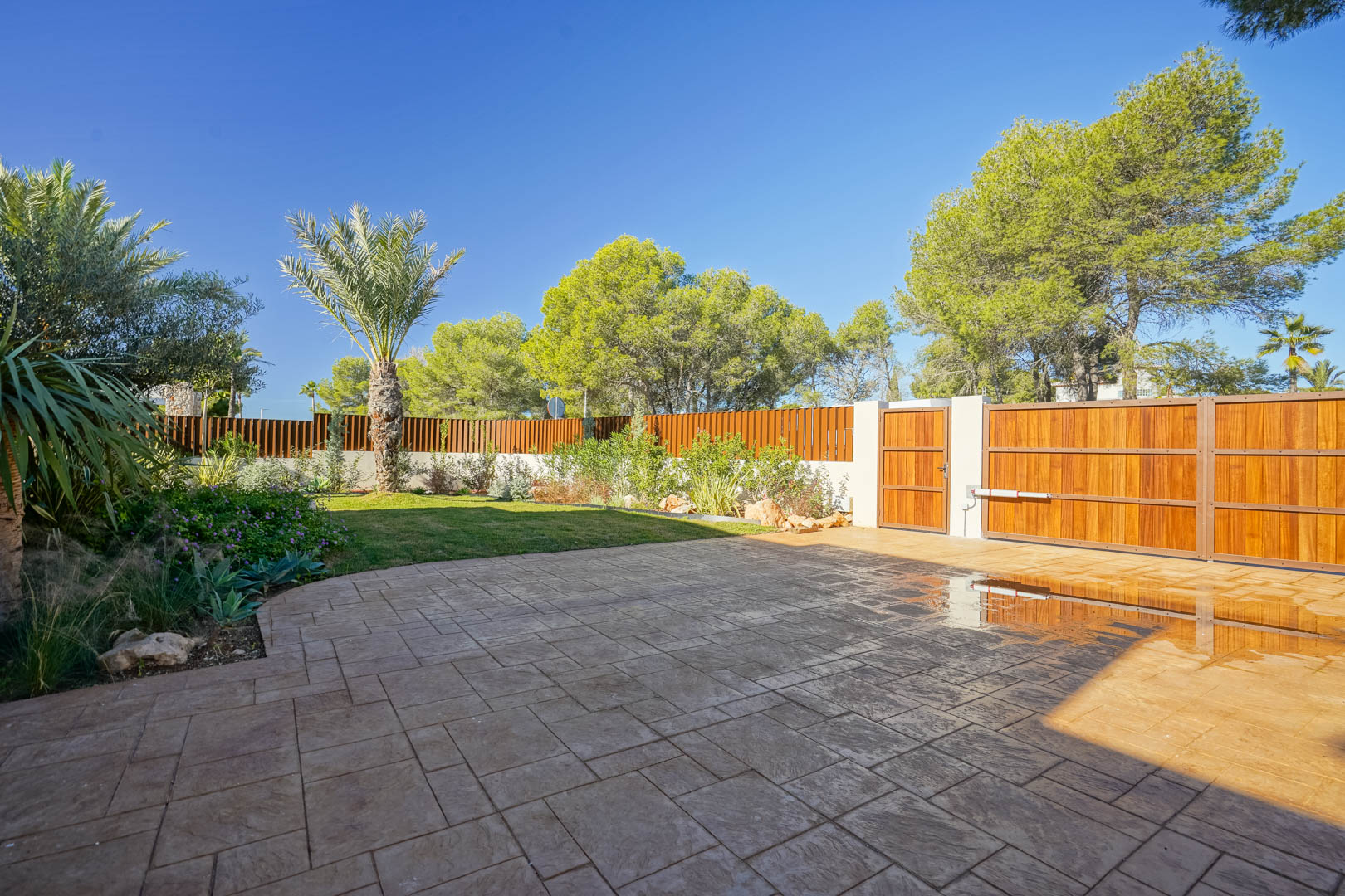 Haupteingang mit motorisiertem Holztor, gepflasterter Einfahrt und mediterranem Garten mit Palmen und Sichtschutzzaun aus Holzlamellen.