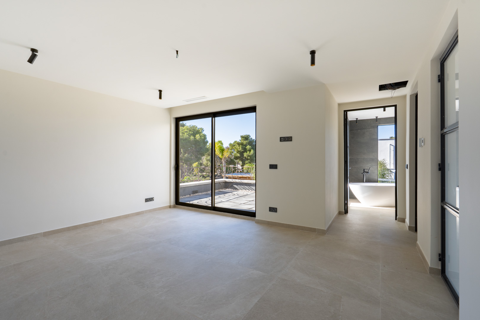 Hauptschlafzimmer mit Zugang zur privaten Terrasse, großformatigen Fliesen und En-Suite-Badezimmer mit freistehender Badewanne und dunklen Wandfliesen.