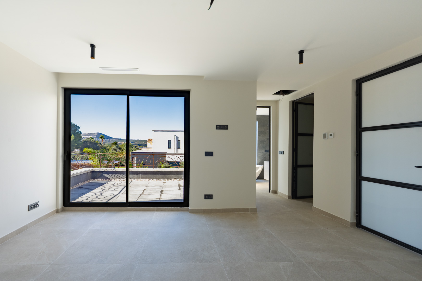 Hauptschlafzimmer mit Zugang zur Terrasse, großformatigen Fliesen und schwarzen Akzenten. Das en-suite Badezimmer mit Badewanne ist im Hintergrund sichtbar.
