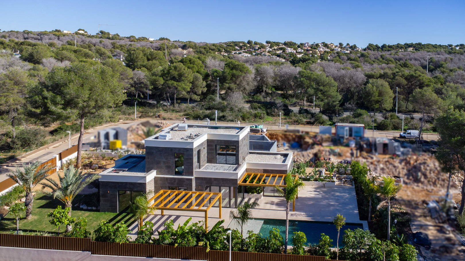 Luftaufnahme der modernen Villa mit dunkler Steinfassade, Iroko-Holzpergolen und Pool, umgeben von einem üppigen mediterranen Garten am Waldrand.