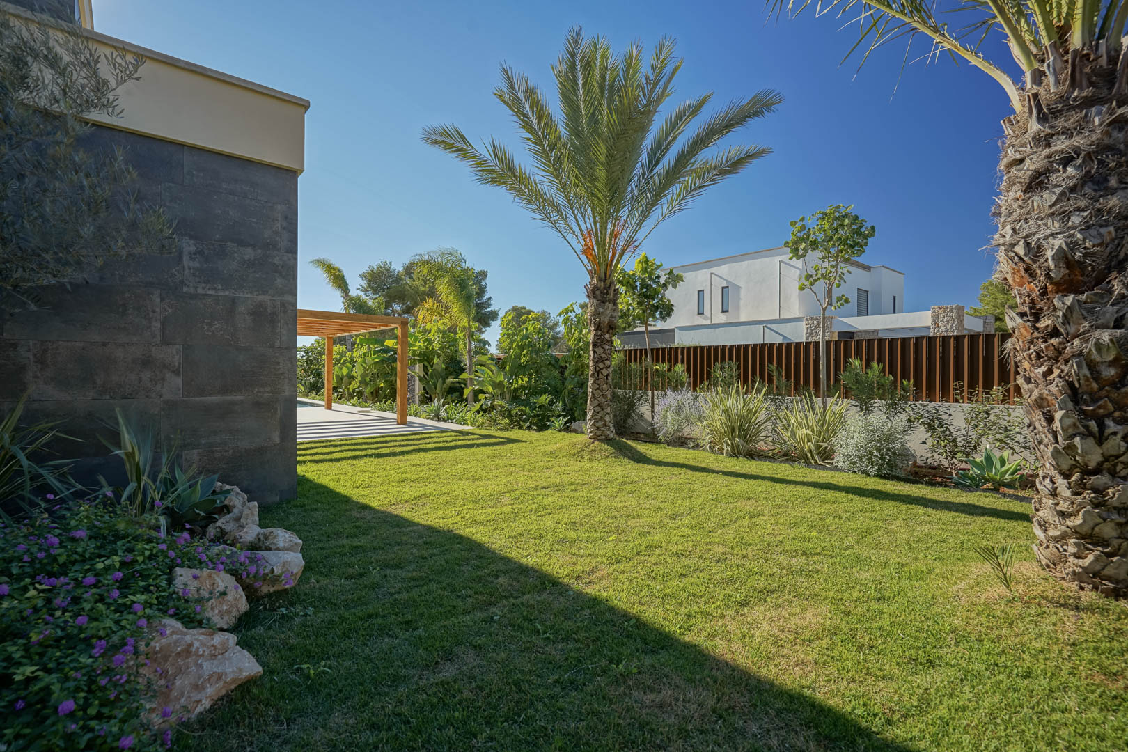 Mediterraner Garten mit Naturrasen, Palmen und einheimischen Pflanzen neben einer dunklen Steinfassade und einer Pergola aus Iroko-Holz.
