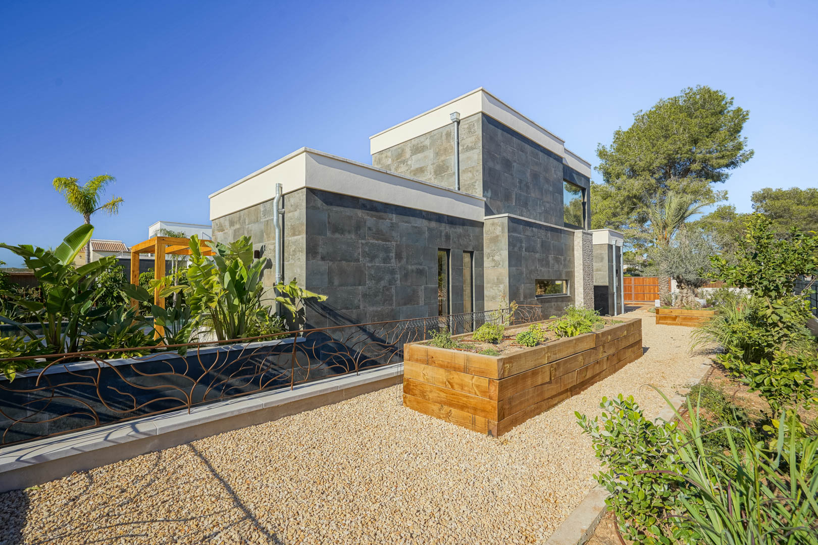 Moderne Fassade mit dunkler Natursteinverkleidung, Hochbeeten aus Holz und Kieswegen in einem gepflegten mediterranen Garten.