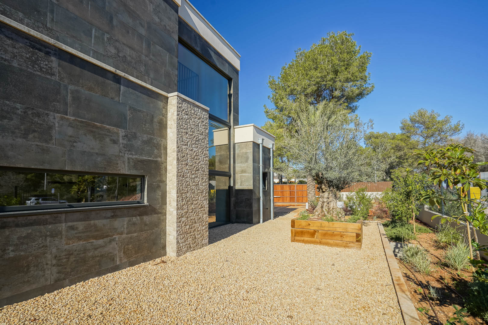 Moderne Fassade mit dunkler Steinverkleidung, strukturierten Natursteinakzenten, großen Glasfronten und mediterranem Garten mit Olivenbaum in Holzhochbeet.