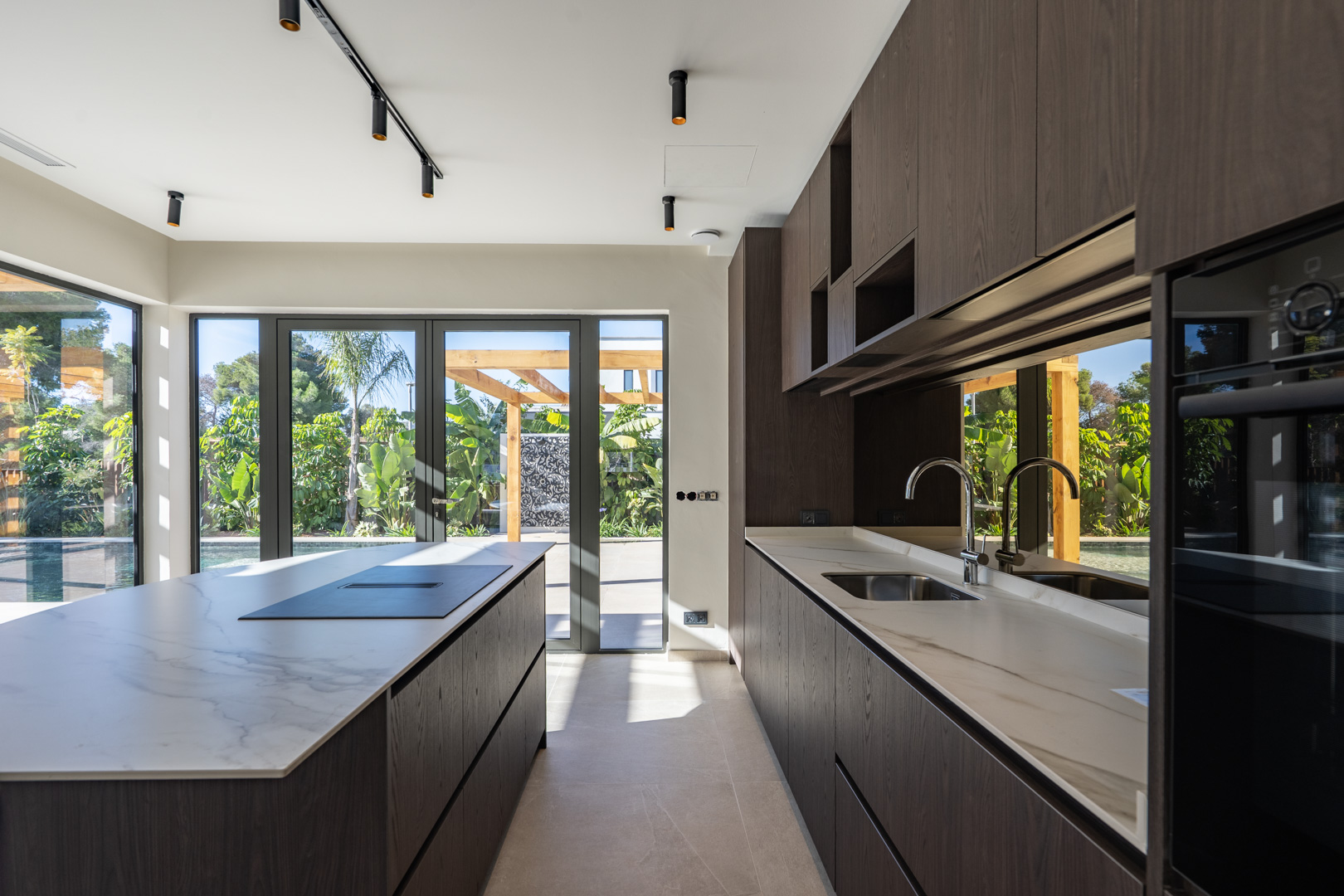 Moderne Küche mit Kochinsel, Arbeitsplatten in Marmoroptik und dunklen Holzfronten. Große Glasfronten bieten direkten Zugang zum Garten und Poolbereich.