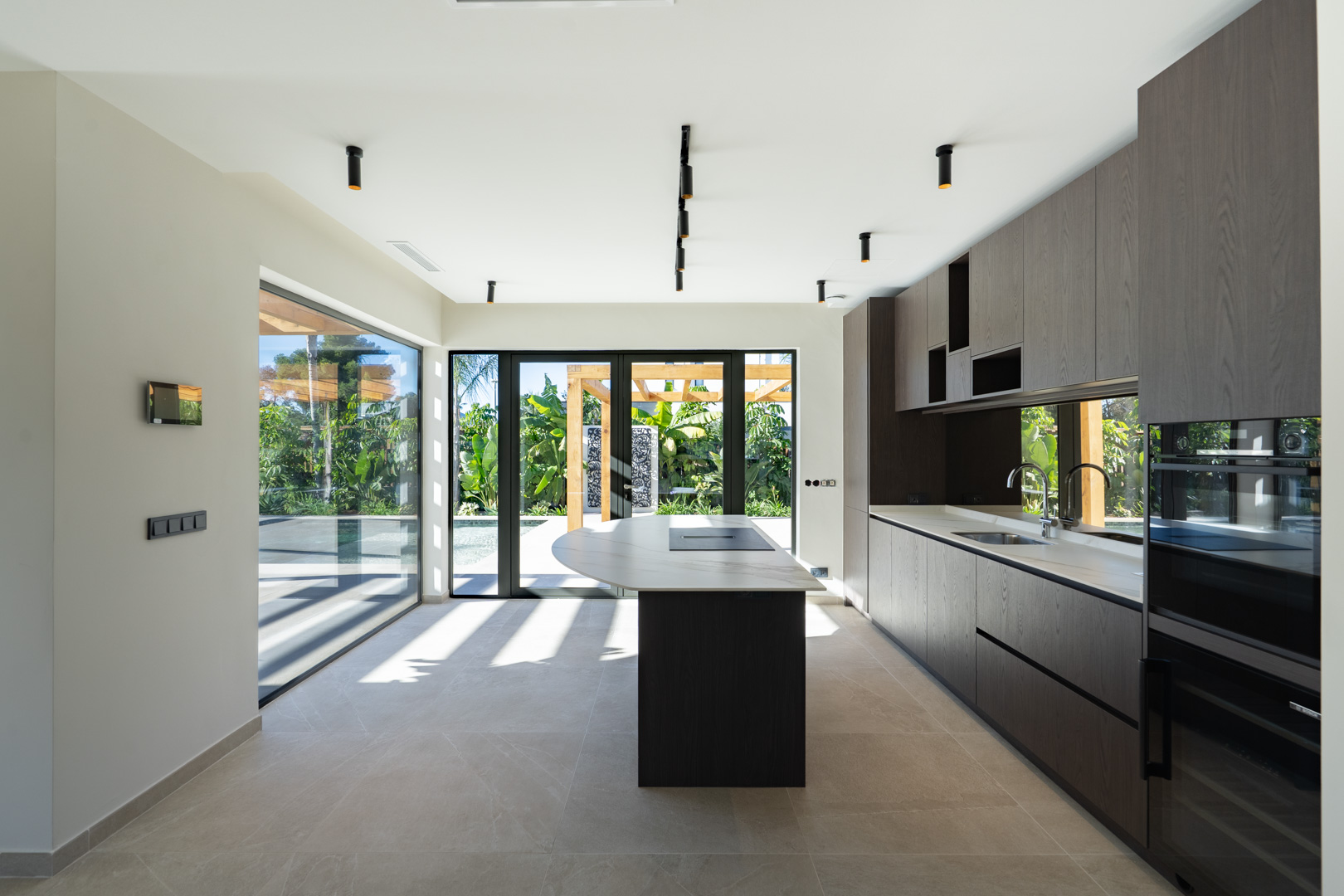 Moderne Küche mit Kochinsel, dunklen Holzfronten und hochwertigen Arbeitsplatten. Große Fensterfronten bieten einen direkten Blick auf den Garten und den Poolbereich.