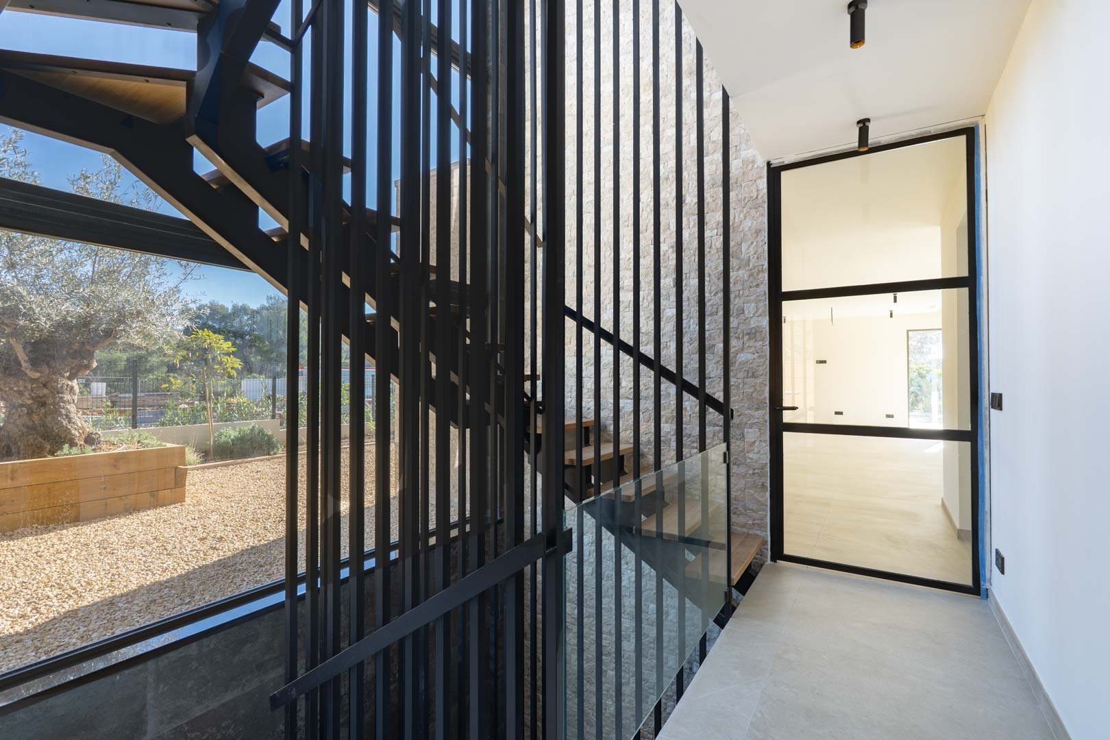 Moderne Treppe mit Holzstufen, schwarzen Metallstreben und einer Natursteinwand, lichtdurchflutet durch große Fenster mit Blick auf den mediterranen Garten.