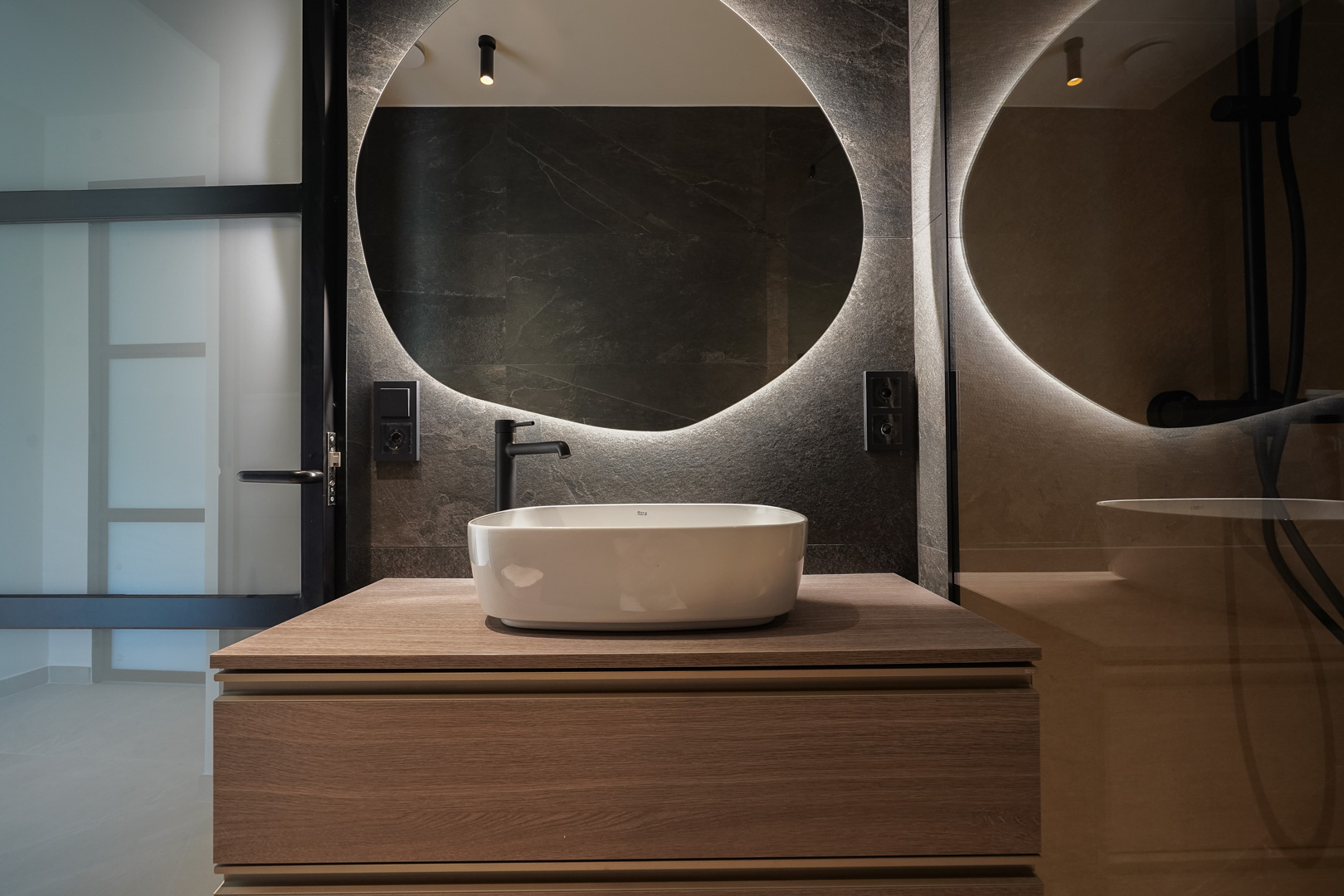 Modernes Bad mit Aufsatzwaschbecken auf Holzmöbel, hinterleuchtetem Rundspiegel und dunklen Fliesen in Steinoptik mit mattschwarzen Armaturen.