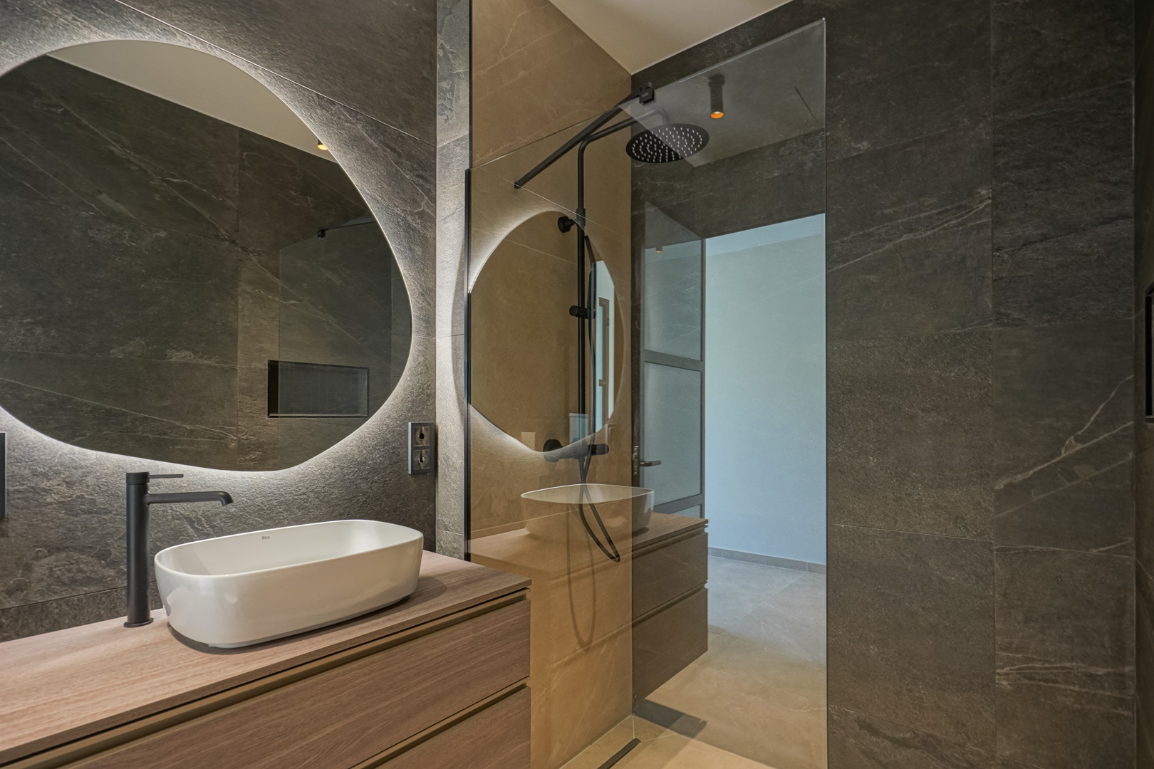 Modernes Badezimmer mit dunklen Fliesen in Steinoptik, Holz-Waschtisch mit Aufsatzbecken und hinterleuchtetem Rundspiegel neben einer begehbaren Dusche.