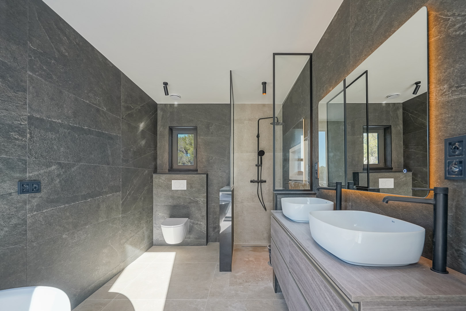 Modernes Badezimmer mit grauen Steinfliesen, Doppelwaschbecken auf Holzunterschrank, mattschwarzen Armaturen und einer begehbaren Dusche mit Glaswand.