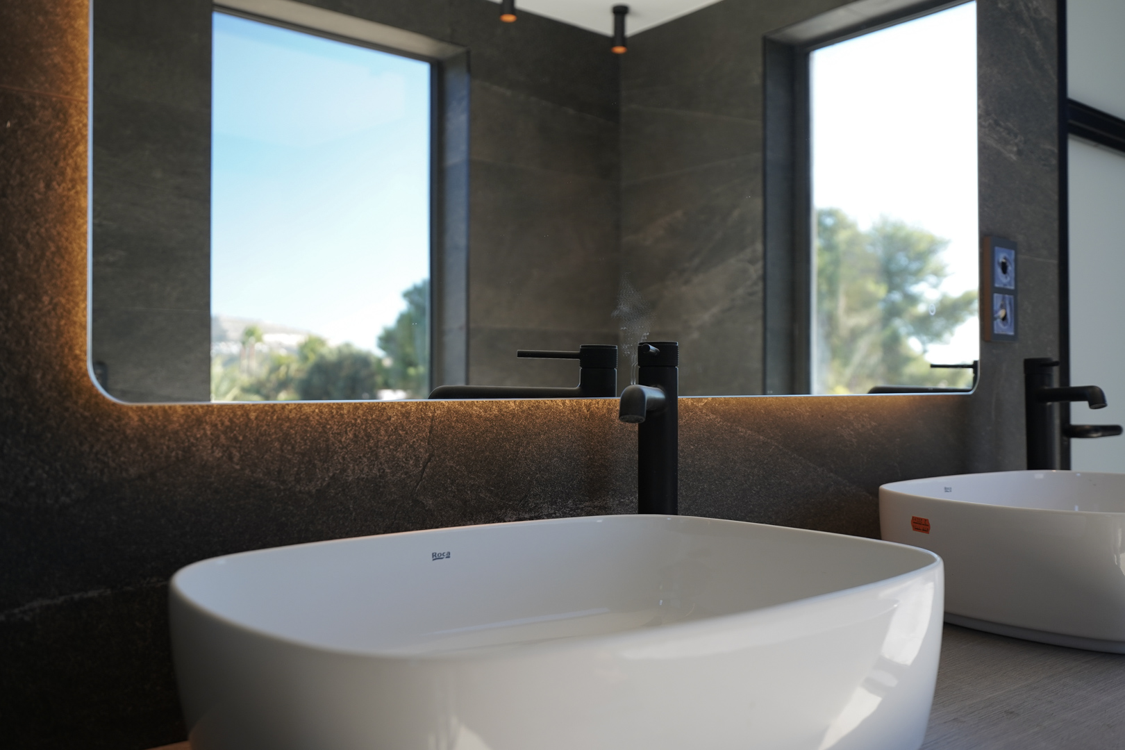 Modernes Badezimmer mit Roca-Aufsatzwaschbecken, mattschwarzen Armaturen und hinterleuchtetem Spiegel vor dunklen Steinwänden.