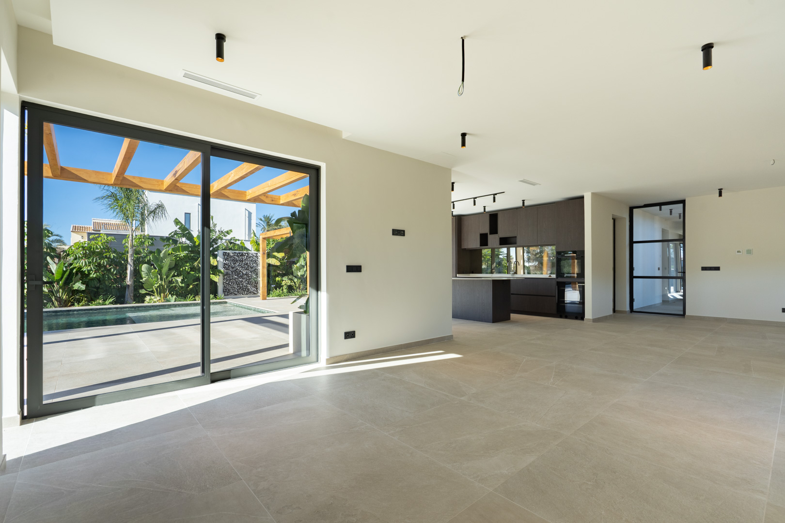 Offener Wohnbereich mit Feinsteinzeugböden und großen Glasschiebetüren zur Terrasse mit Pool und Iroko-Pergola. Im Hintergrund befindet sich die moderne dunkle Holzküche.