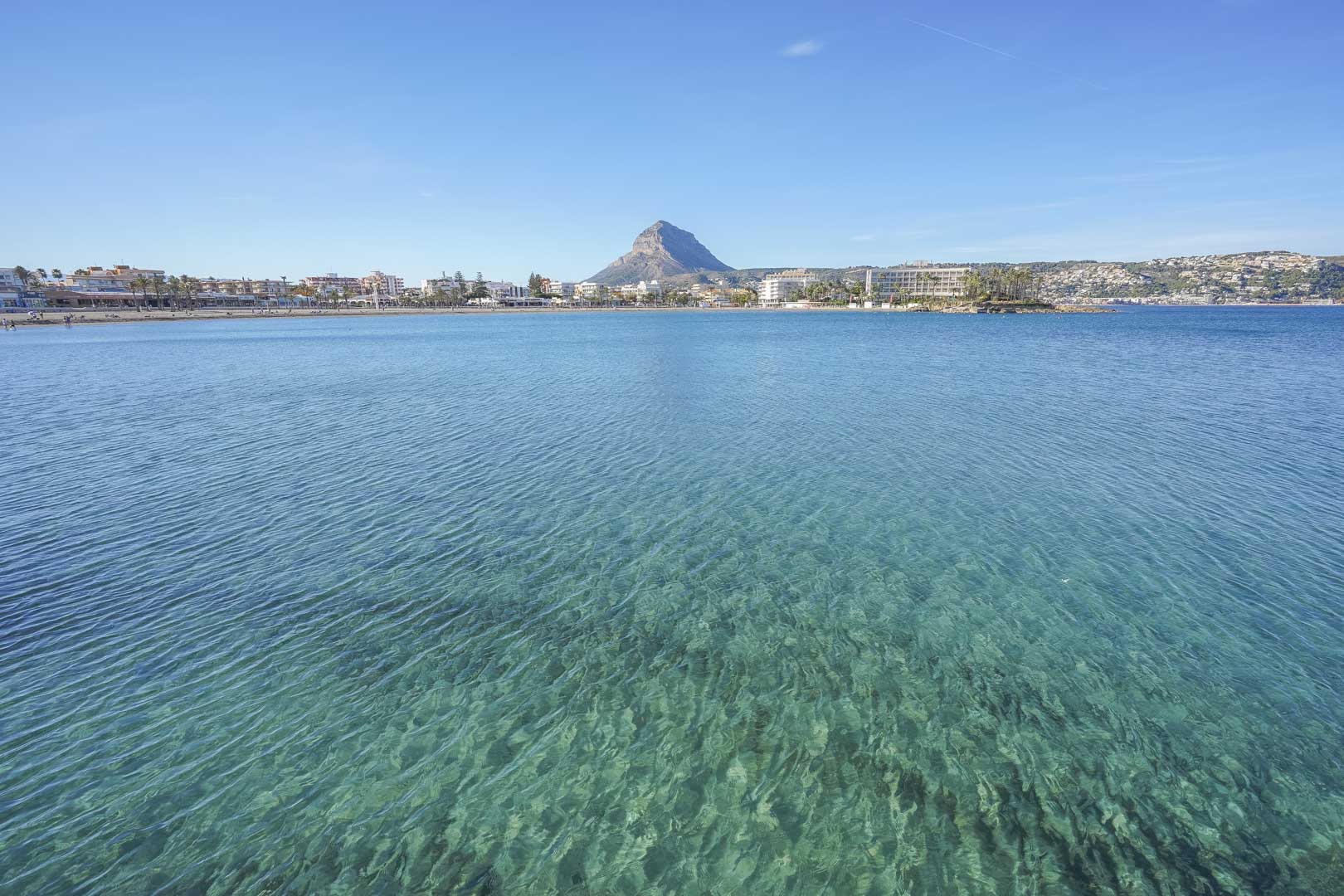 Panoramablick auf die Bucht von Jávea mit kristallklarem, türkisblauem Wasser und dem markanten Berg Montgó im Hintergrund.
