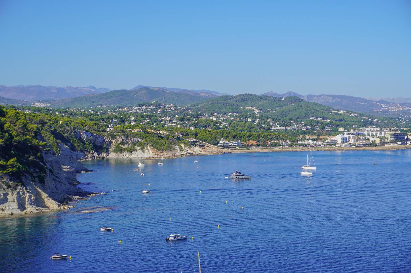 Panoramablick auf die Küste von Jávea mit ankernden Booten im blauen Wasser, mediterranen Klippen und Bergen im Hintergrund bei klarem Himmel.