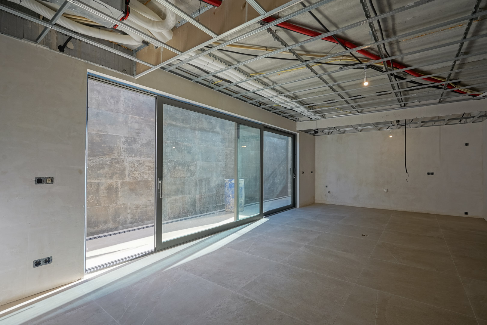 Rohbau des Untergeschosses mit großen Glasschiebetüren, großformatigen Bodenfliesen und sichtbaren Deckeninstallationen für Lüftung und Elektrik.
