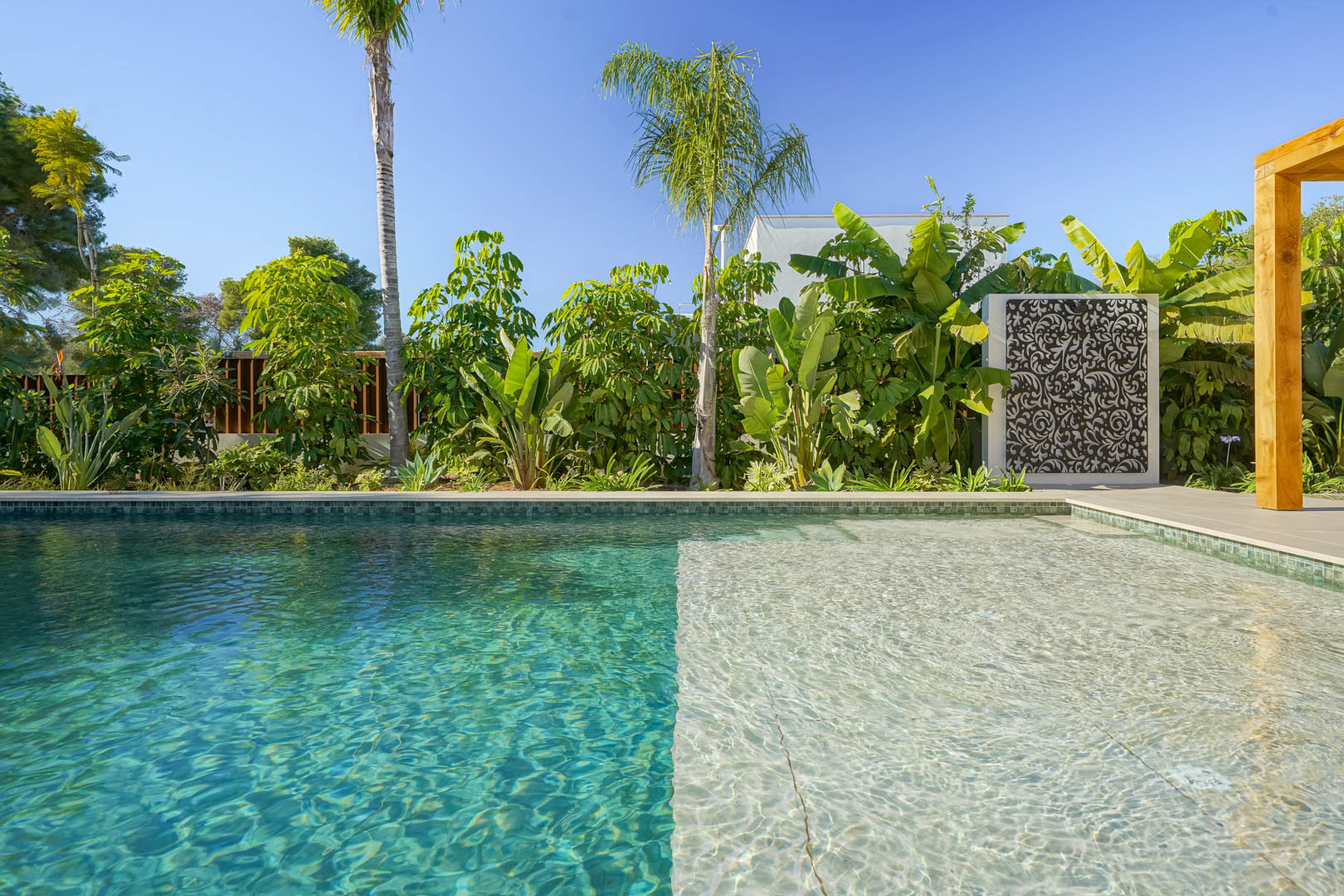 Salzwasserpool im Bali-Stil mit Flachwasserzone, umgeben von einem üppigen mediterranen Garten mit Palmen und einer dekorativen Trennwand.