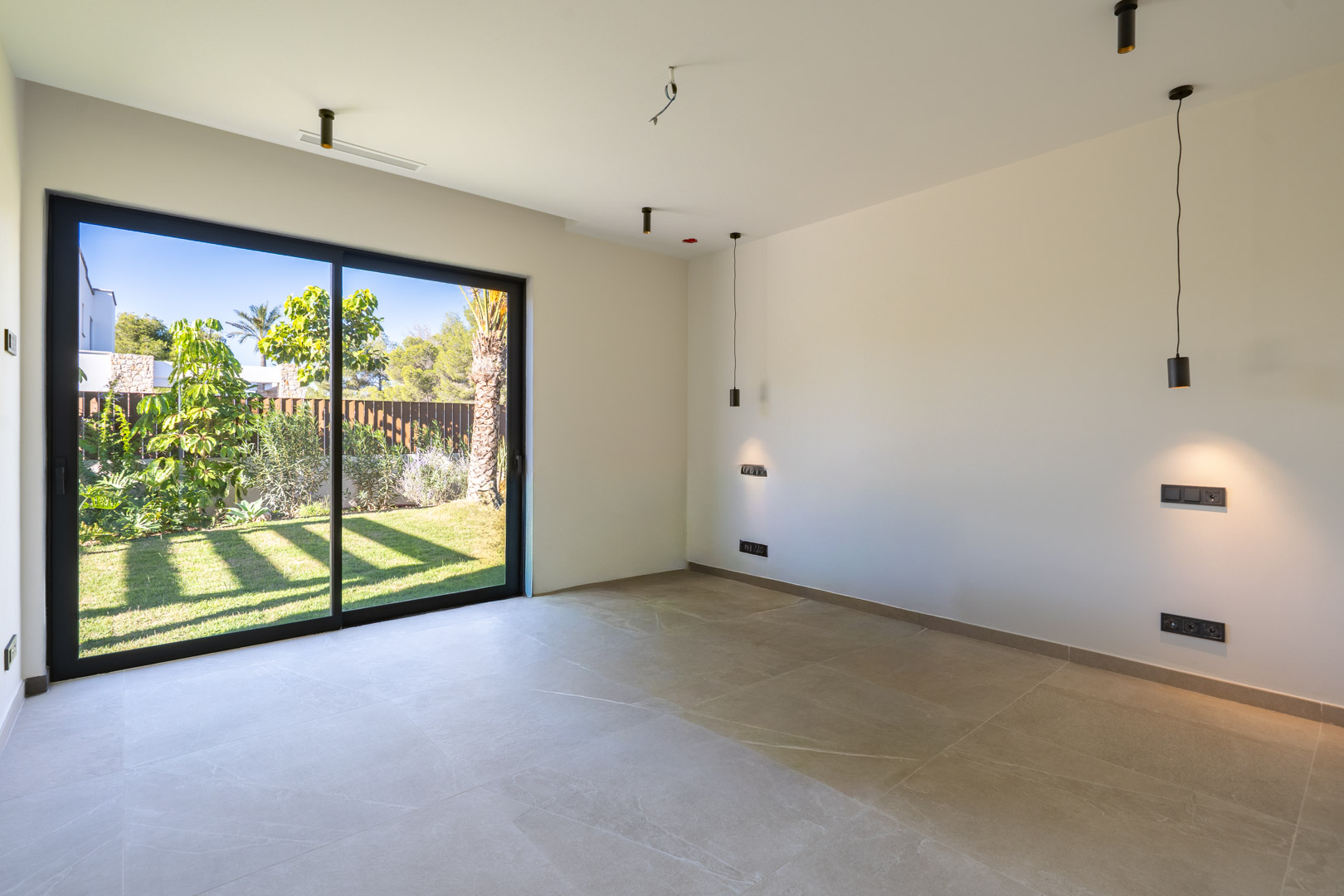 Schlafzimmer mit Feinsteinzeugfliesen und großen Glasschiebetüren zum Garten. Ausgestattet mit minimalistischen schwarzen Pendelleuchten und integrierter Klimaanlage.