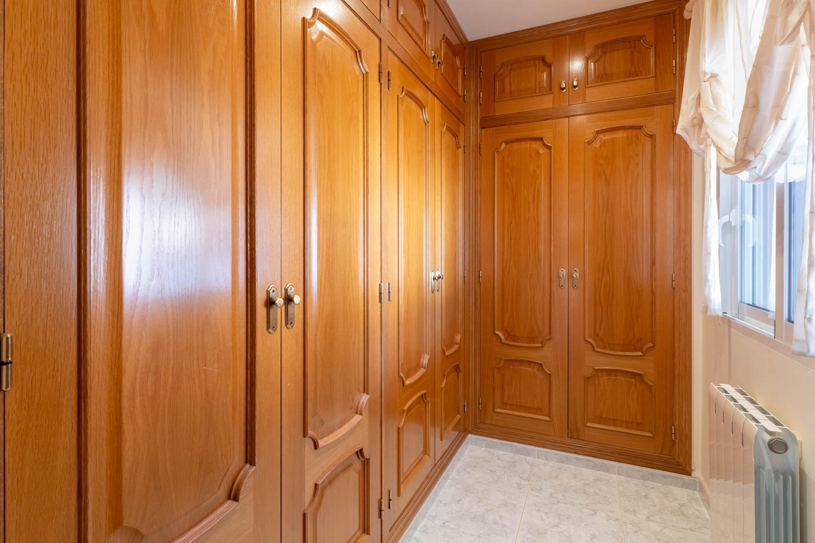 Ankleidezimmer mit großzügigen Einbauschränken aus Holz, klassischen Paneeltüren, Fliesenboden und einem Heizkörper unter dem Fenster.