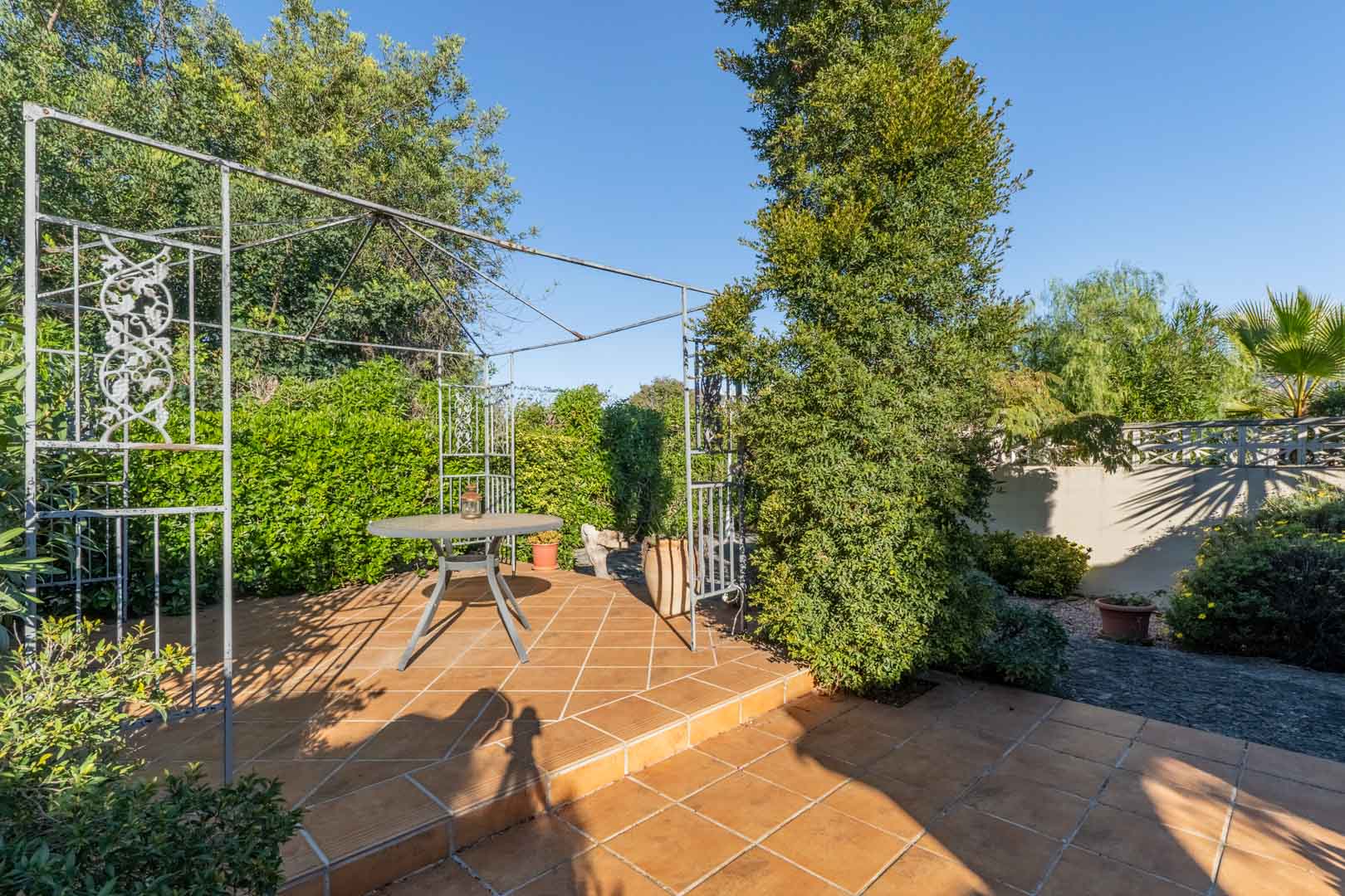 Außenbereich mit Terrakotta-Fliesen, einer dekorativen Metallpergola und einem runden Tisch, umgeben von üppiger mediterraner Bepflanzung und Hecken.