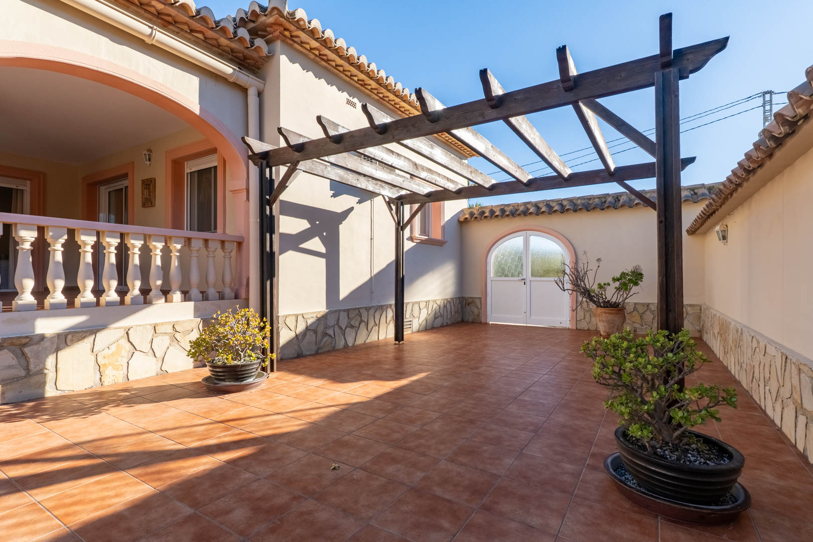 Außenbereich mit Terrakotta-Fliesen, einer Holzpergola und Natursteinsockel. Die Terrasse führt zur erhöhten Naya mit klassischer Balustrade.
