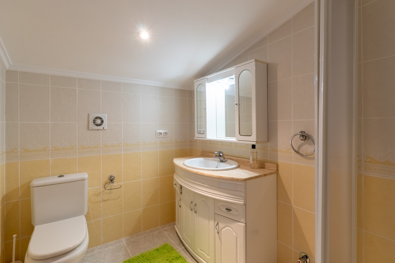 Badezimmer mit creme- und gelbfarbenen Fliesen, geschwungenem Waschtisch mit Marmorplatte und Spiegelschrank mit integrierter Beleuchtung.