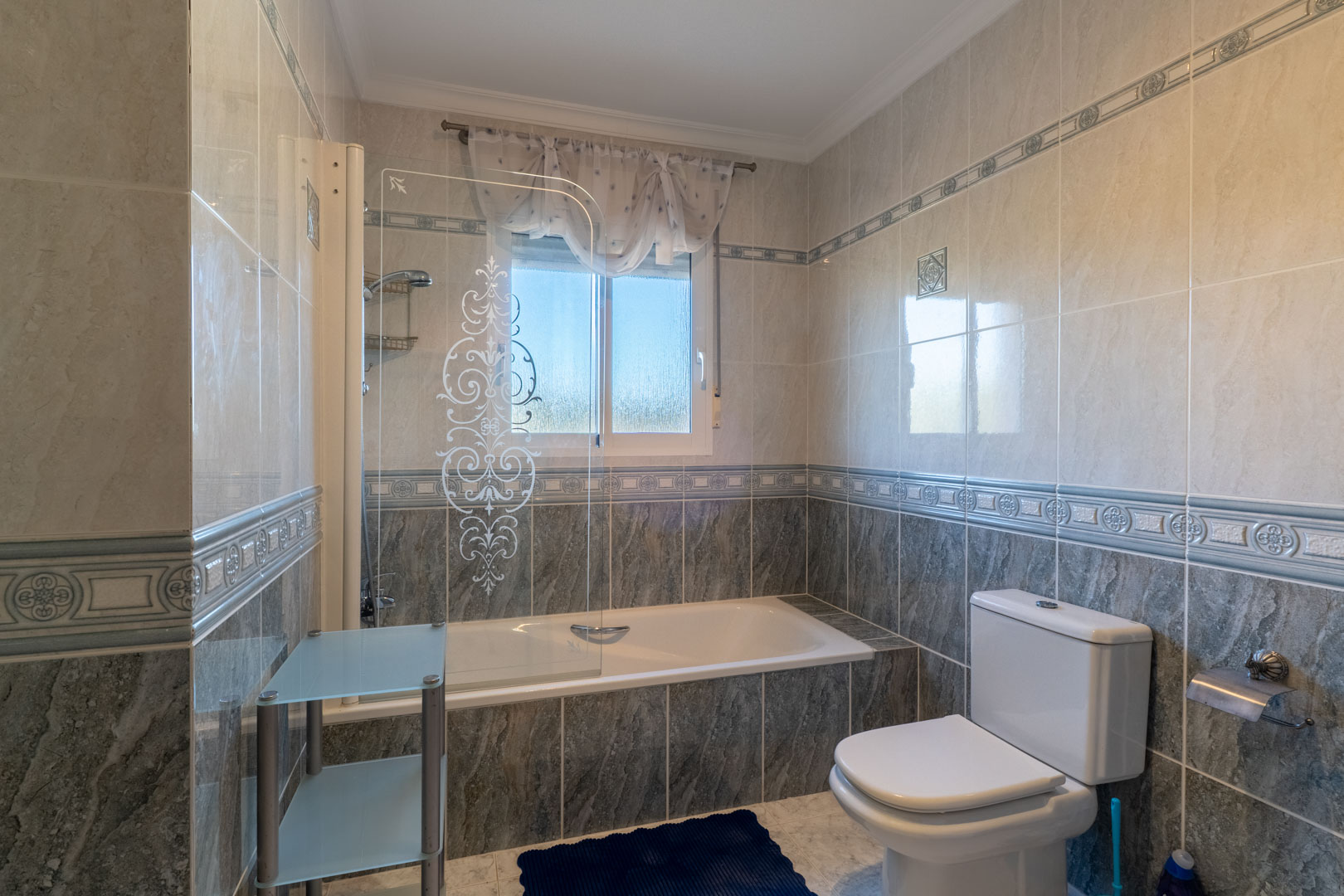Badezimmer mit Einbaubadewanne, dekorierter Glastrennwand und zweifarbigen Fliesen. Ein Fenster sorgt für natürliches Licht und Belüftung.