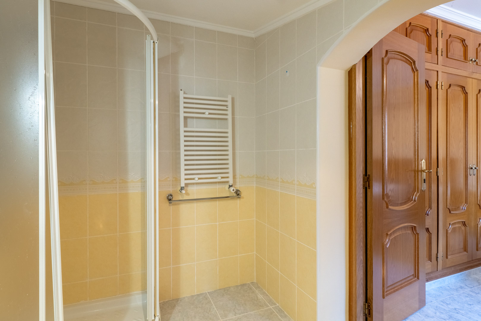 Badezimmer mit zweifarbigen Fliesen, Handtuchheizkörper und Dusche, durch einen Rundbogen mit dem Schlafzimmer und klassischen Holzeinbauschränken verbunden.