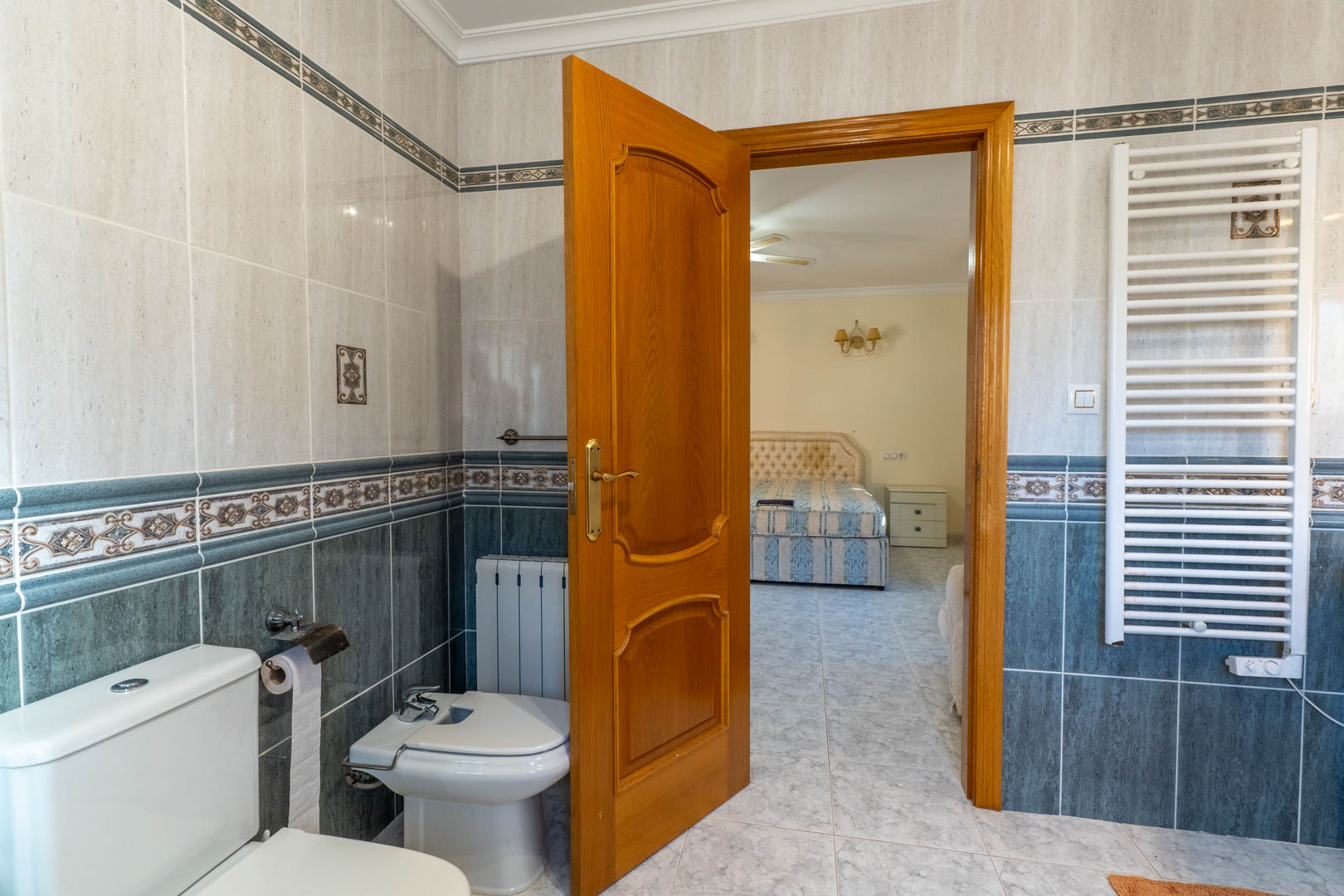 Blick aus dem En-Suite-Badezimmer in das Schlafzimmer, mit dekorativen zweifarbigen Fliesen, weißem Handtuchheizkörper und massiver Holztür.