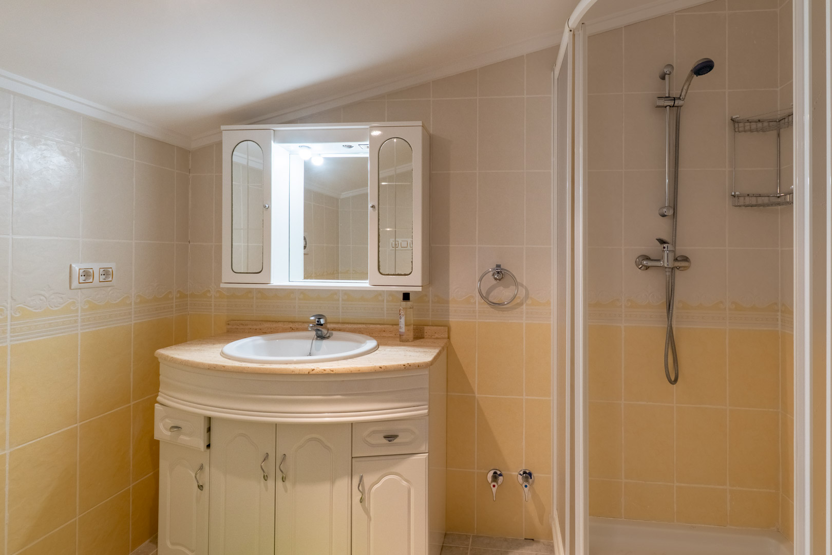 Funktionelles Badezimmer mit Marmorwaschtisch, weißem Unterschrank und Dusche, gestaltet mit cremefarbenen Fliesen und einem beleuchteten Spiegelschrank.