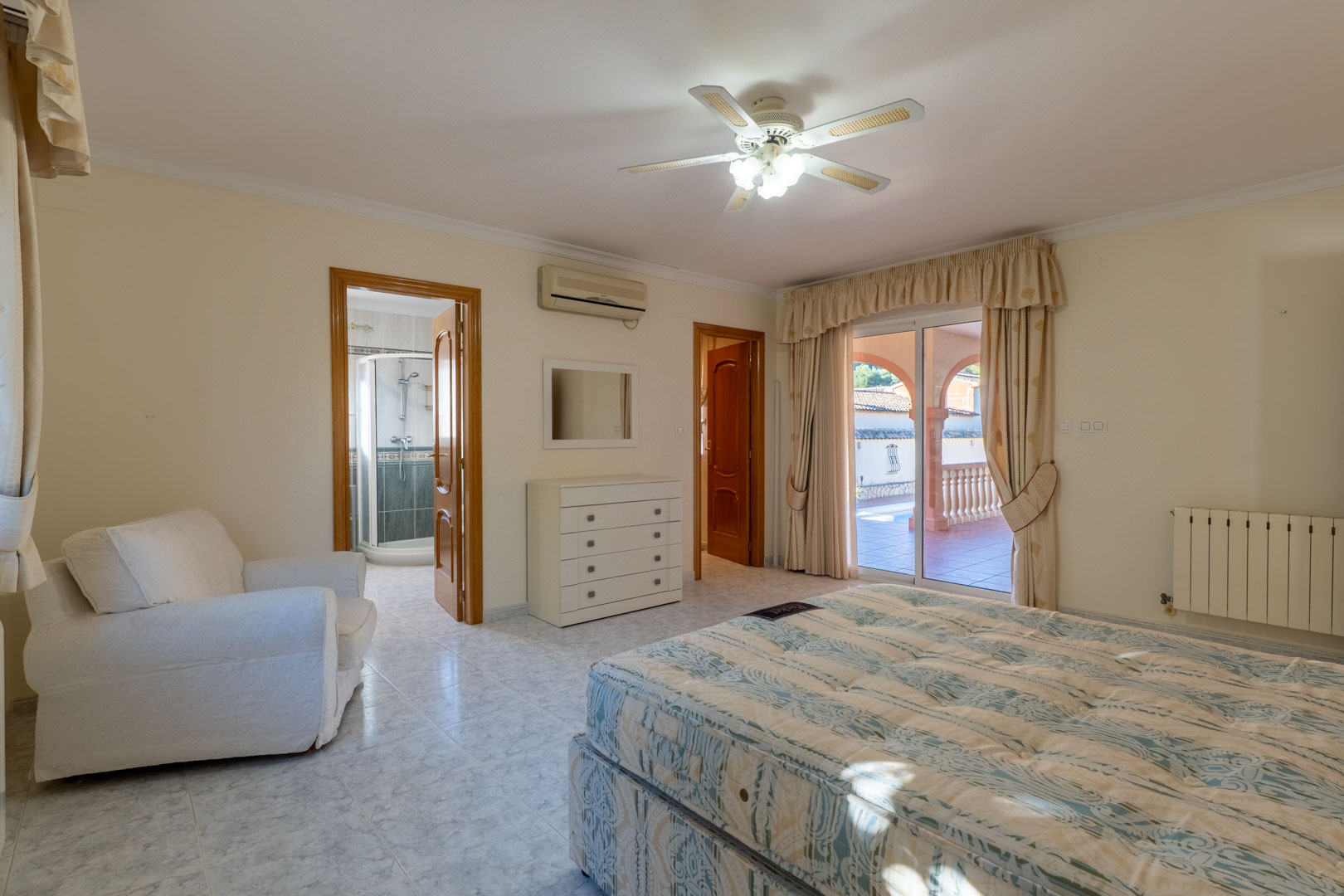 Hauptschlafzimmer mit En-Suite-Bad, Klimaanlage und direktem Terrassenzugang. Es bietet helle Fliesenböden, einen Deckenventilator und Blick auf den Poolbereich.