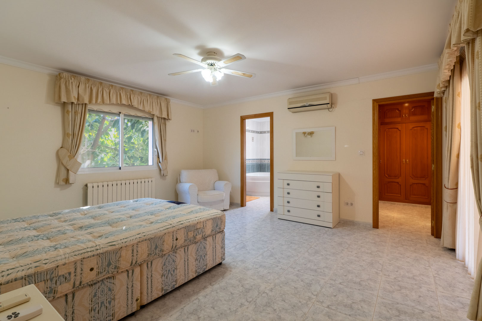 Hauptschlafzimmer mit En-Suite-Badezimmer und begehbarem Kleiderschrank. Ausgestattet mit Fliesenboden, Klimaanlage, Deckenventilator und hellen Wänden mit Holztüren.